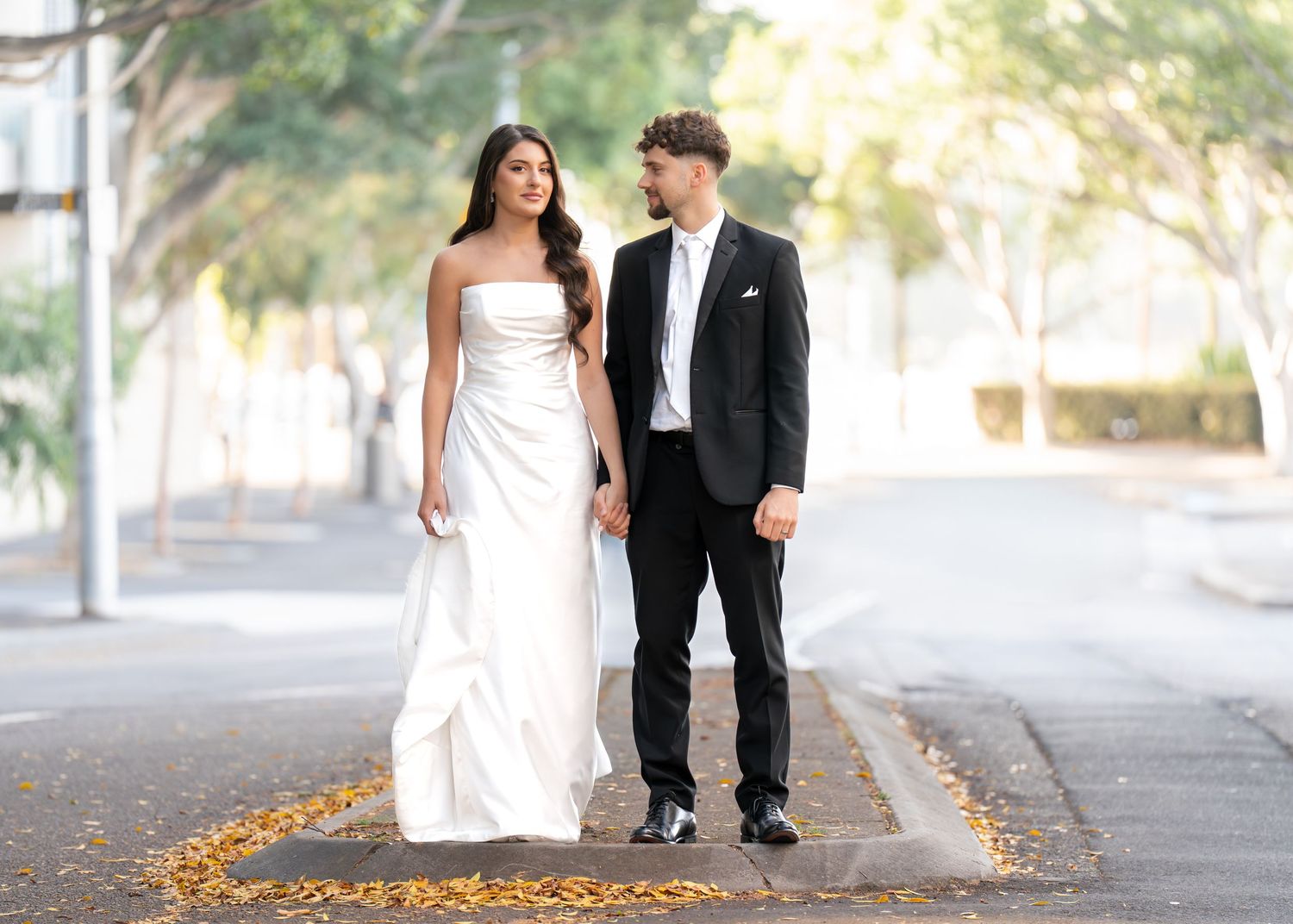 A couple in wedding attire walks along a tree-lined path with autumn leaves scattered on the ground.