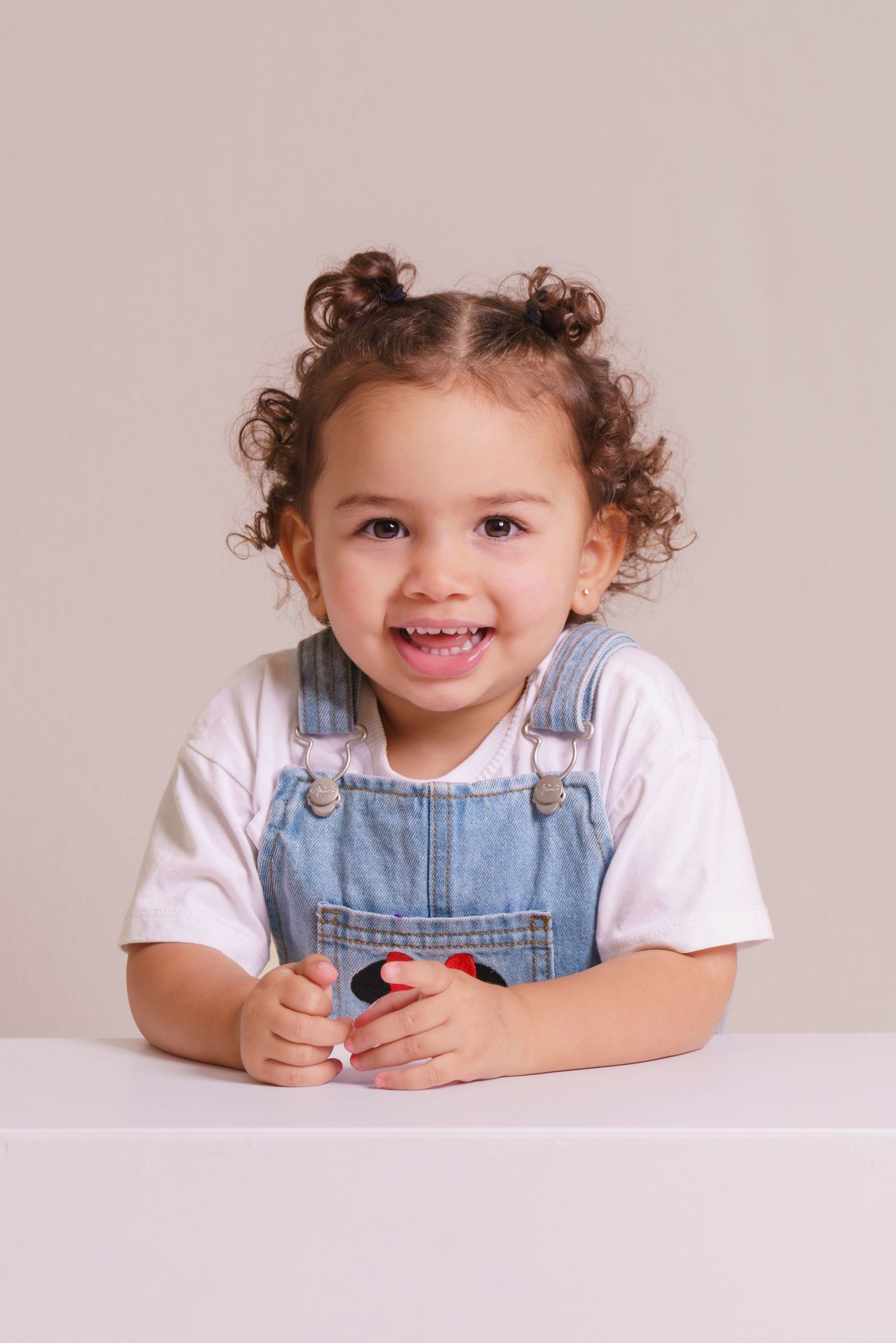 Smiling toddler girl with curly hair in denim overalls and white shirt sitting at a white table.