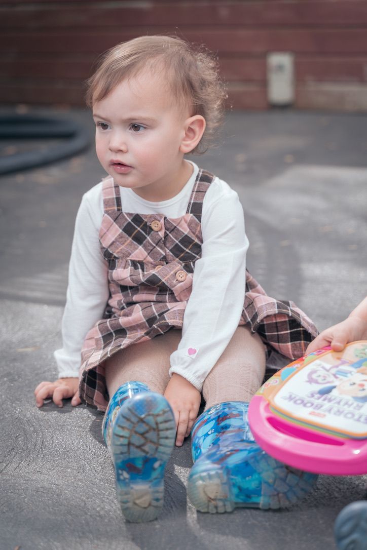 Child in plaid dress and blue rain boots sits on ground with toys.