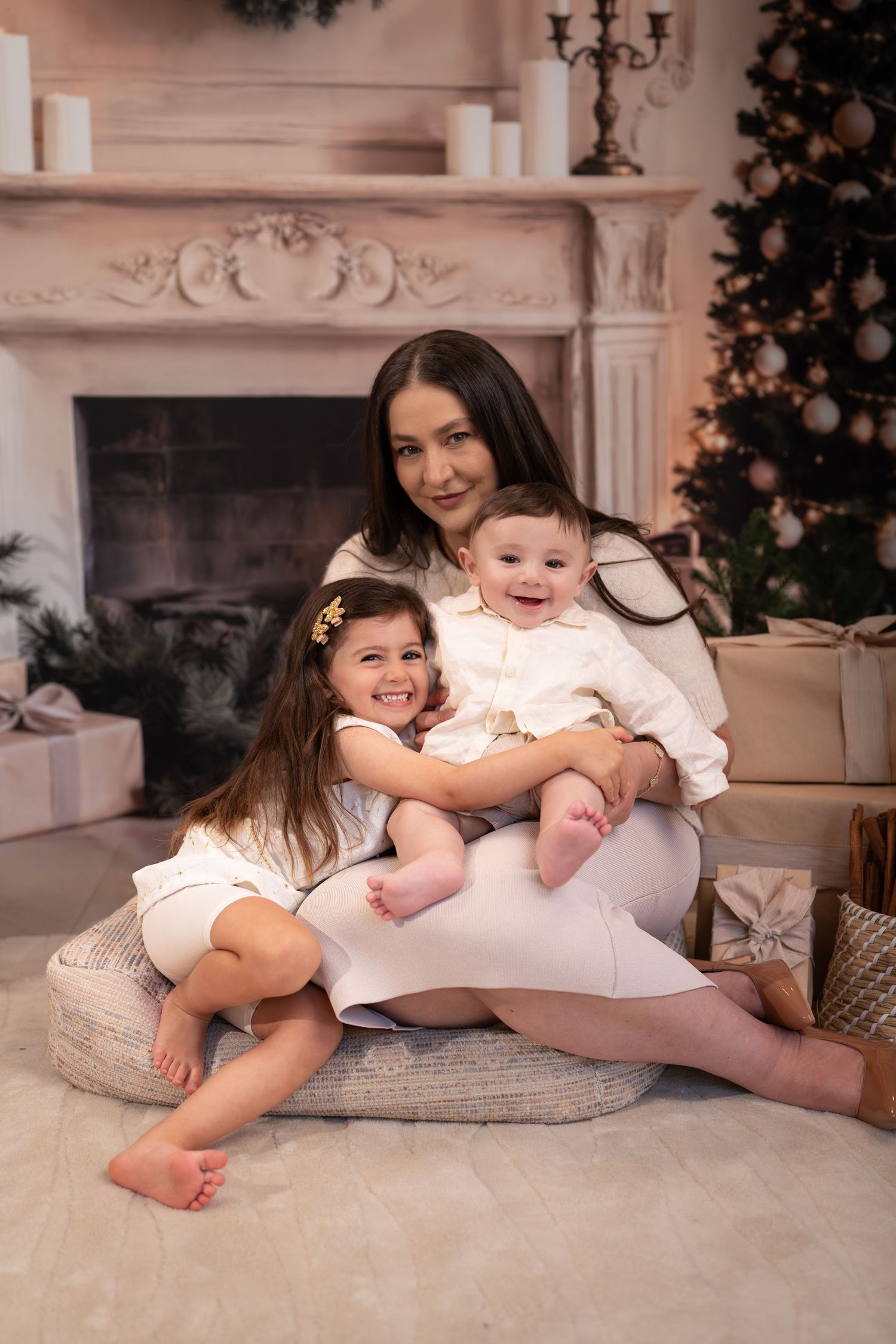 Two young children in white outfits lay on a plush cushion near a decorated fireplace during a holiday photoshoot.