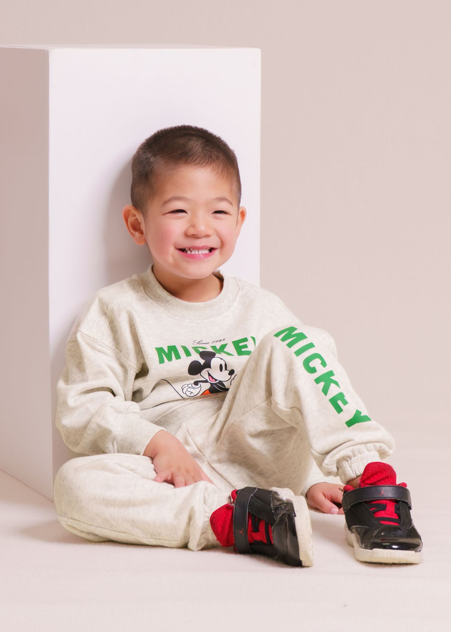 Smiling young boy wearing a Mickey Mouse sweatsuit and black sneakers, sitting against a white backdrop.