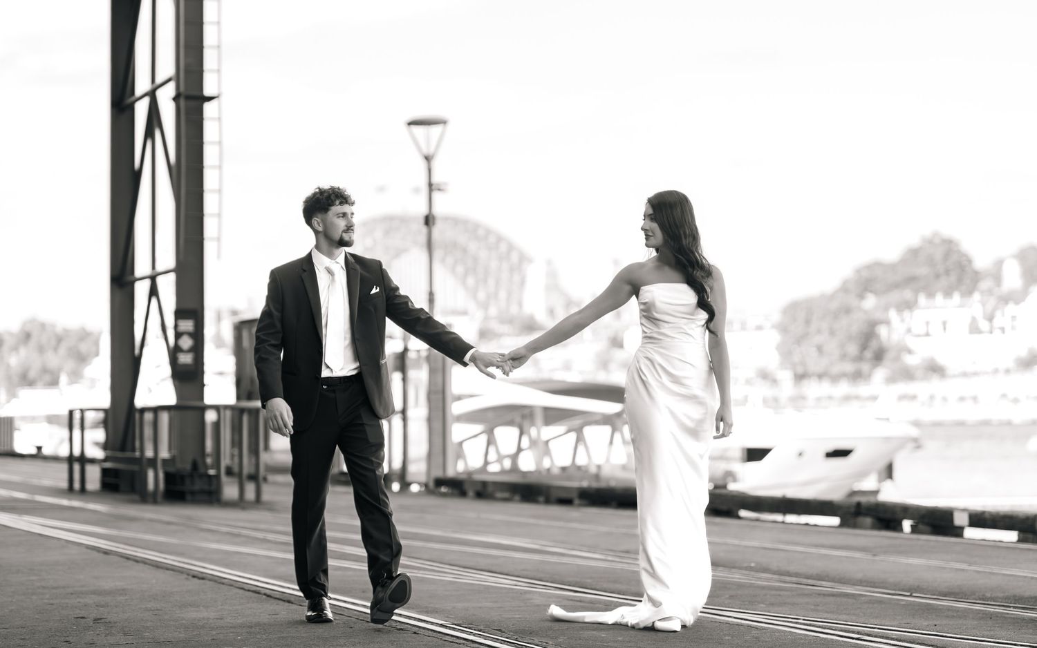 A couple in wedding attire walking hand in hand along a pier with ocean views in the background.