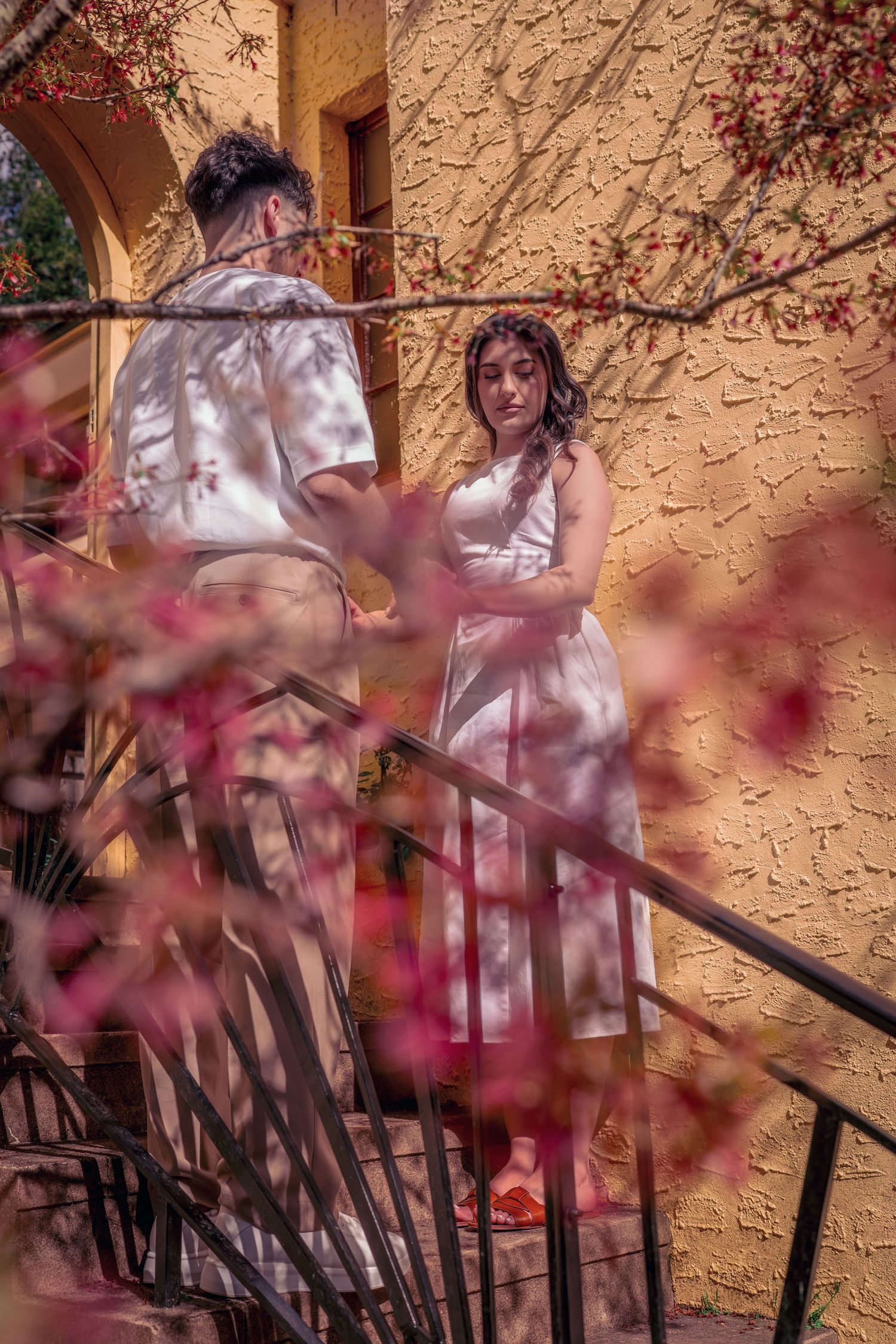 A bride in white dress stands on stairs behind pink cherry blossoms against a textured beige wall.