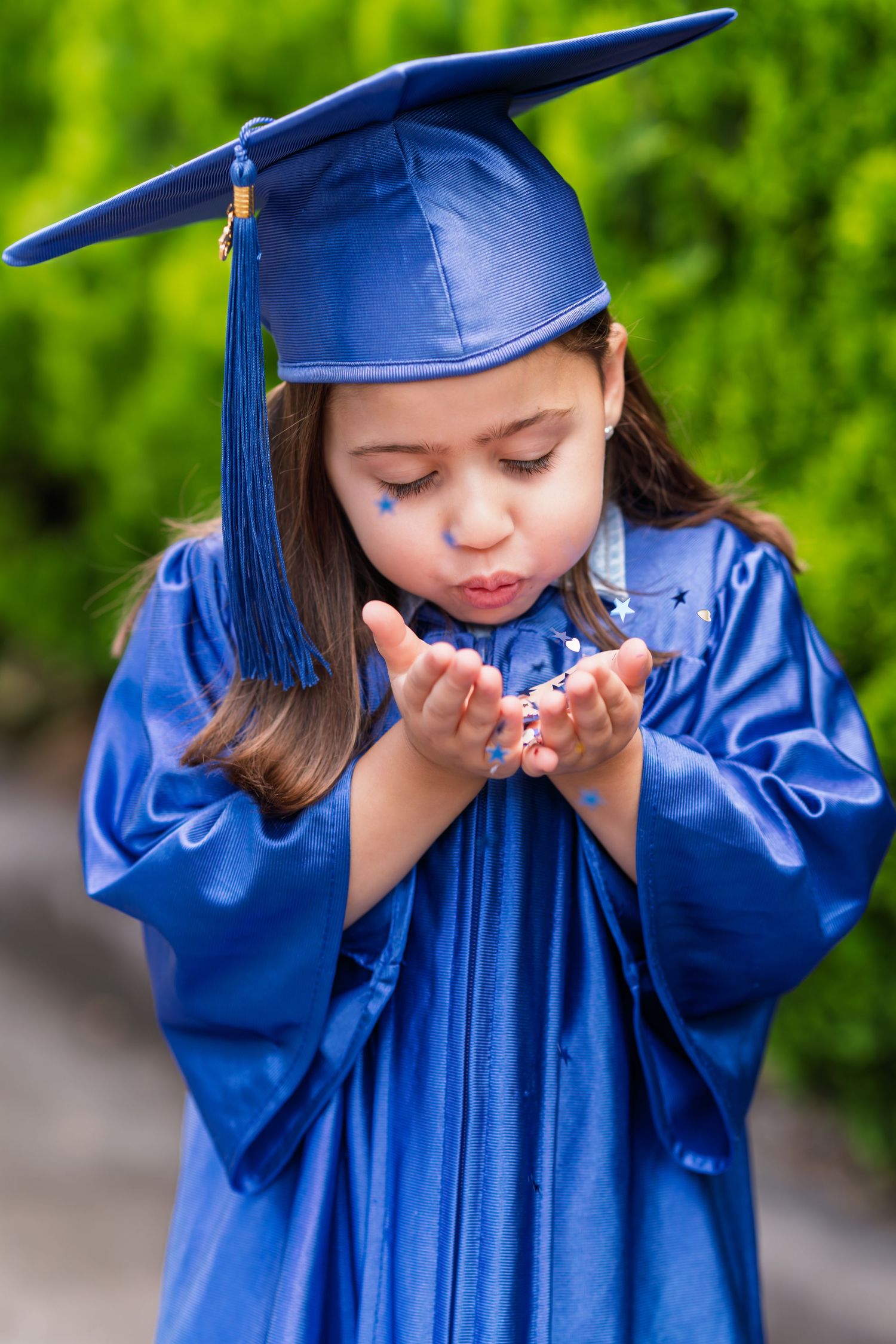 Young graduate in shiny blue cap and gown blows confetti from cupped hands during graduation celebration outdoors.