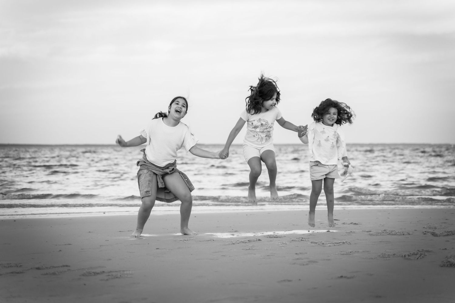 Three figures joyfully jumping and playing together on a beach at sunset in black and white.