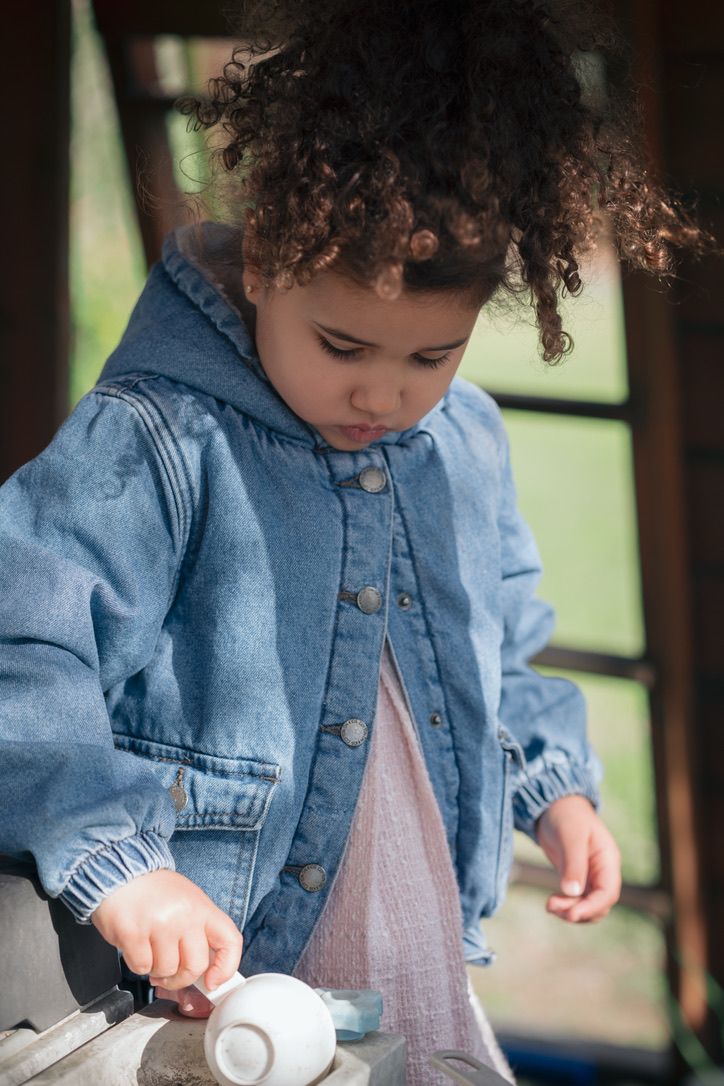 Small child in jean jacket engaged in pouring activity at wooden counter.