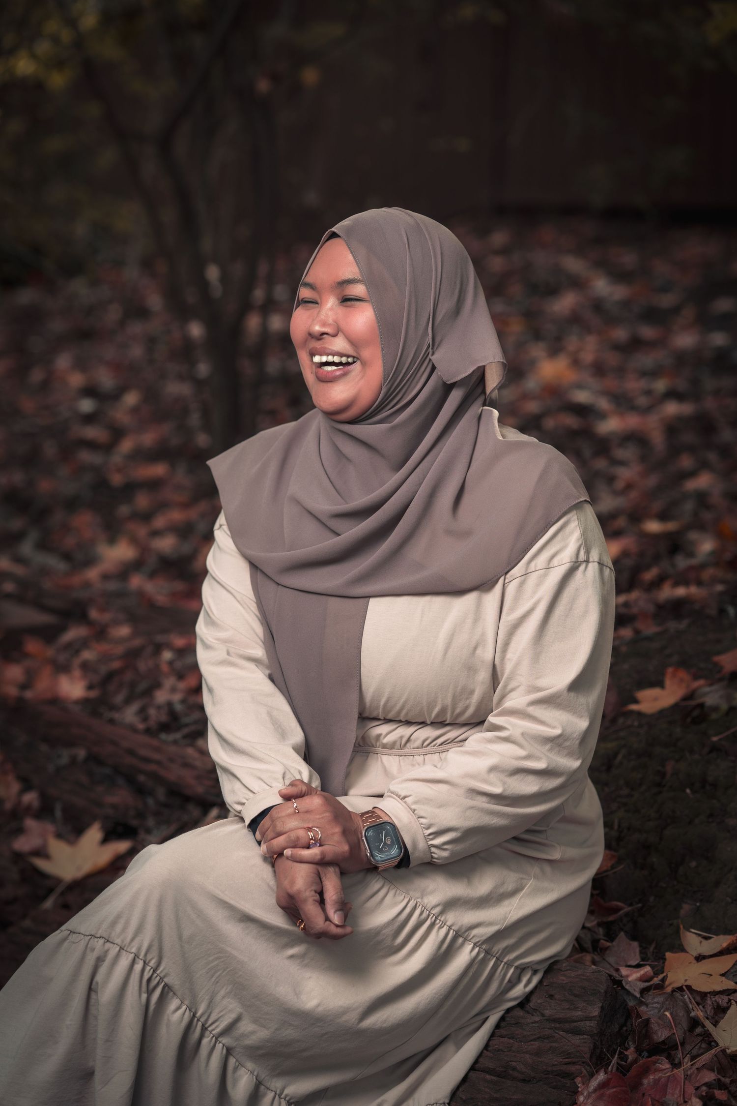 A person in hijab and light dress sitting on autumn leaves, laughing joyfully.