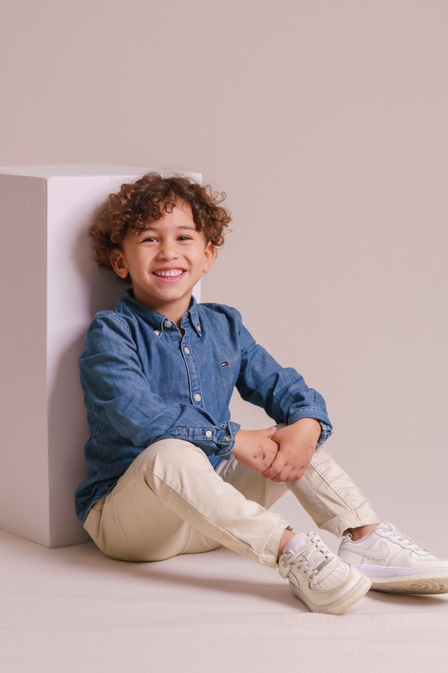 Happy curly-haired boy in a blue denim shirt and beige pants sitting and smiling against a neutral backdrop.