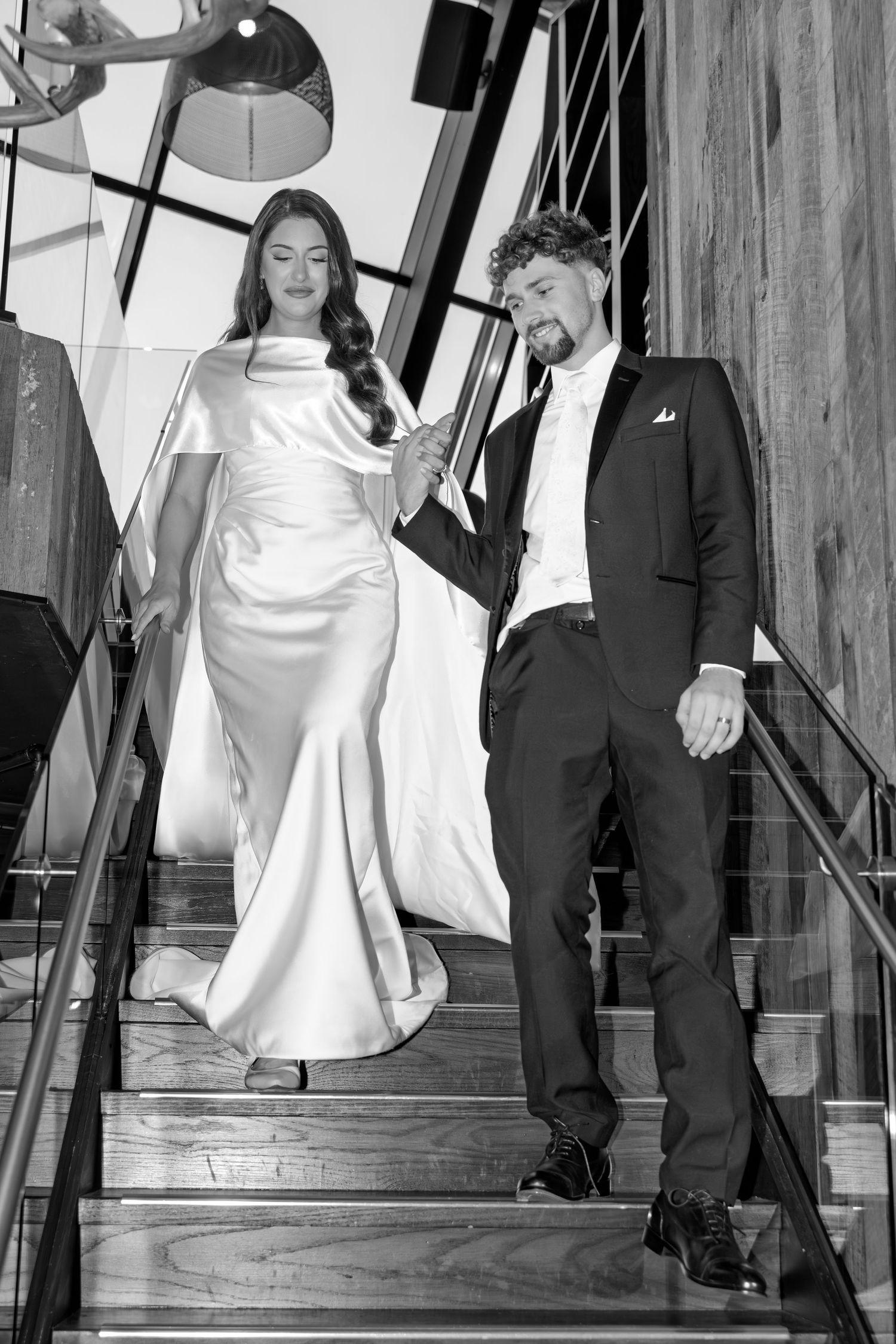 A couple descends a wooden staircase in formal wedding attire shown in black and white.