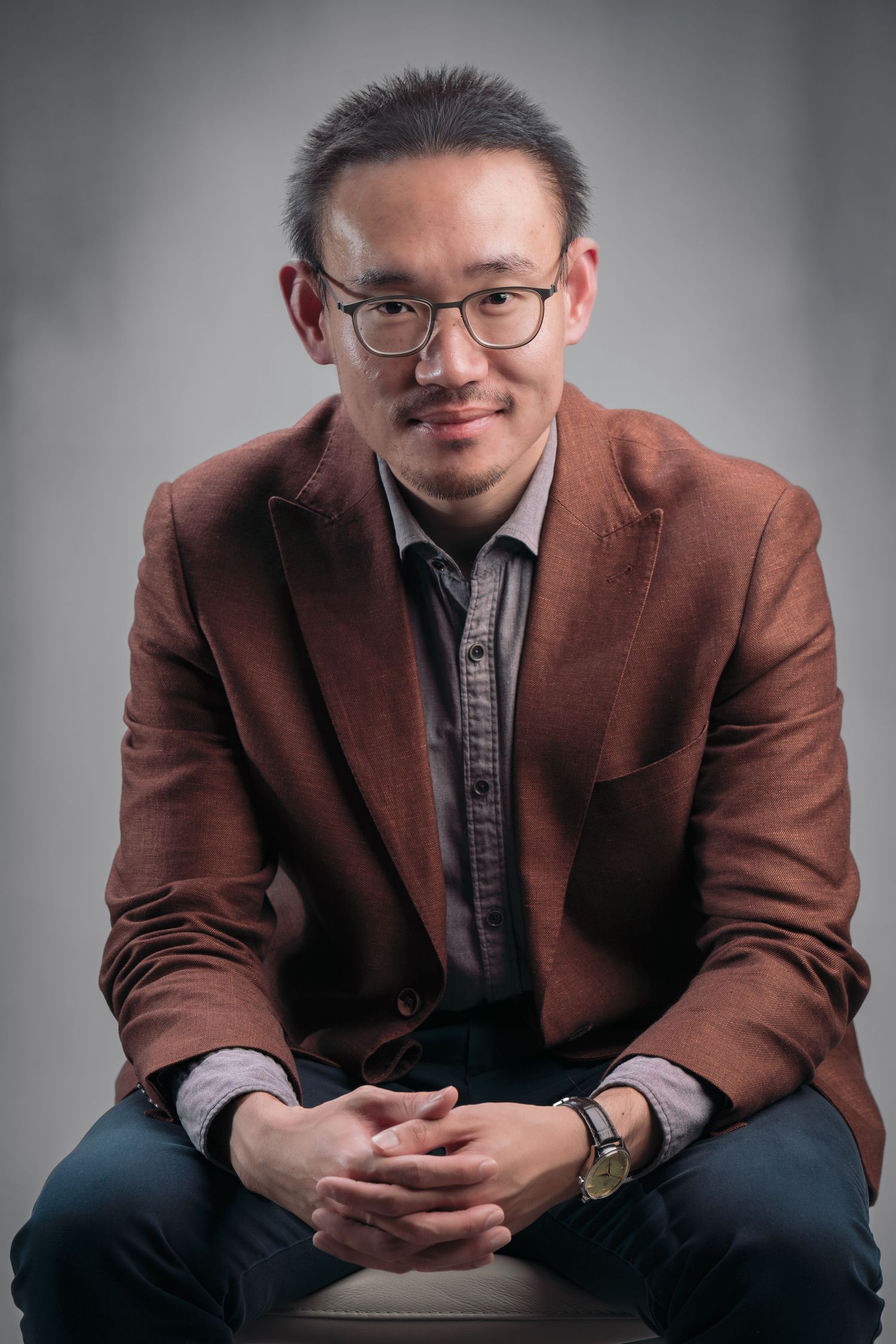 A person wearing glasses and a brown blazer sits casually in a series of portrait poses against a gray background.
