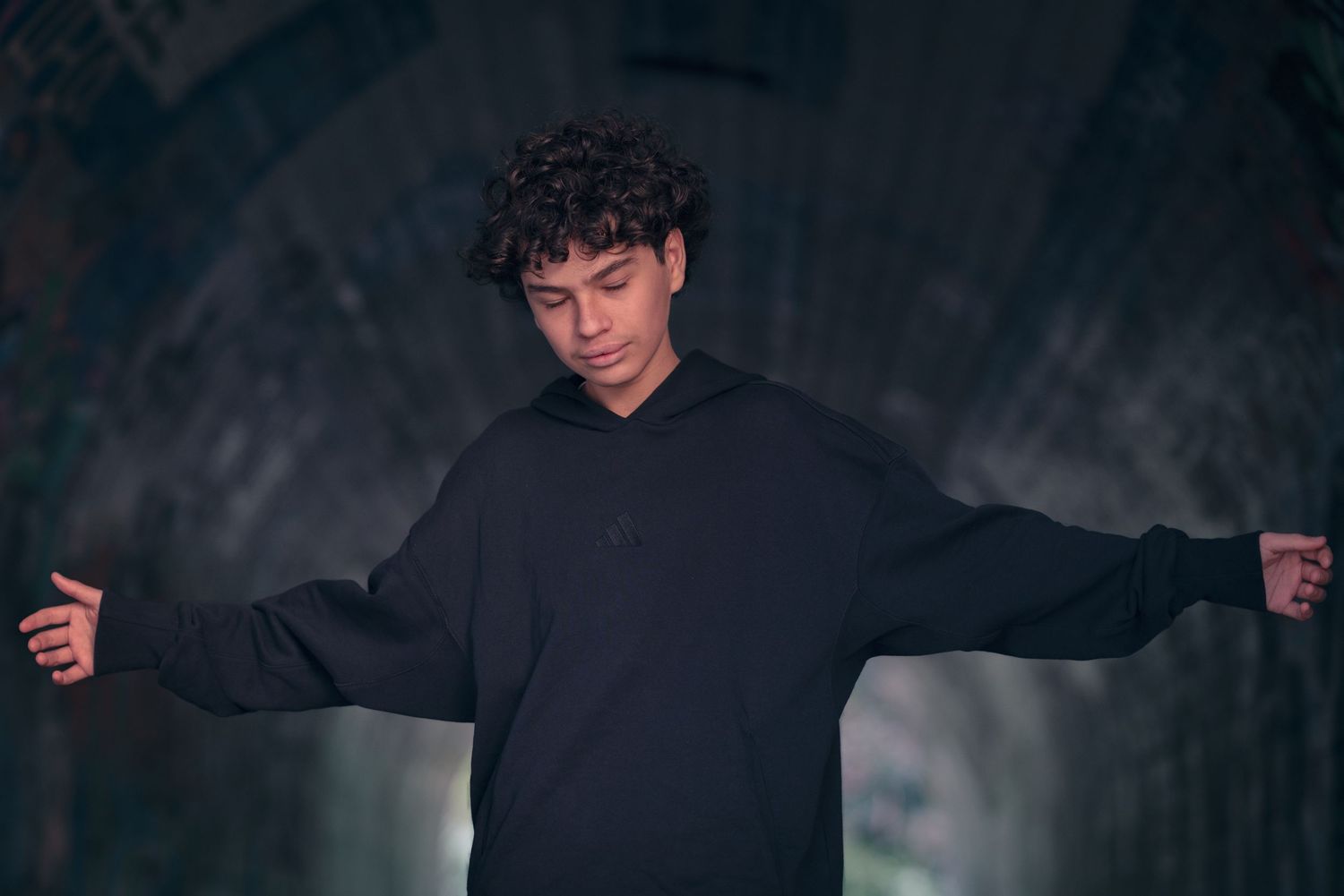 Person in black hoodie with curly hair posing with arms outstretched against dark background.