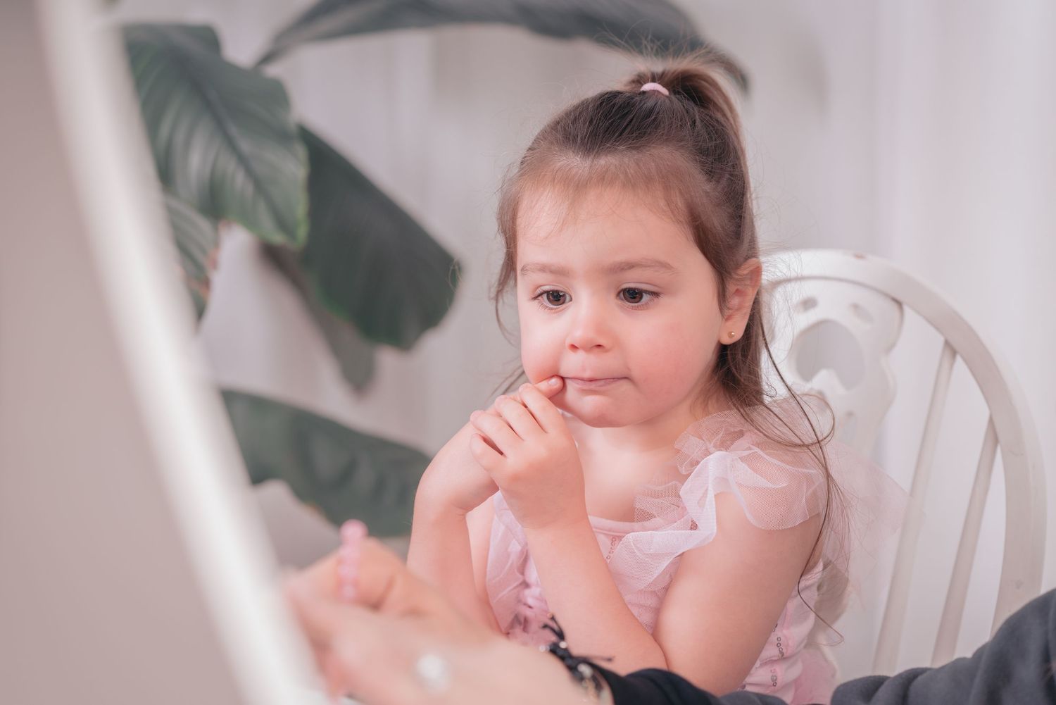 A small child in a pink dress sits thoughtfully in a white chair with a houseplant visible in the background.