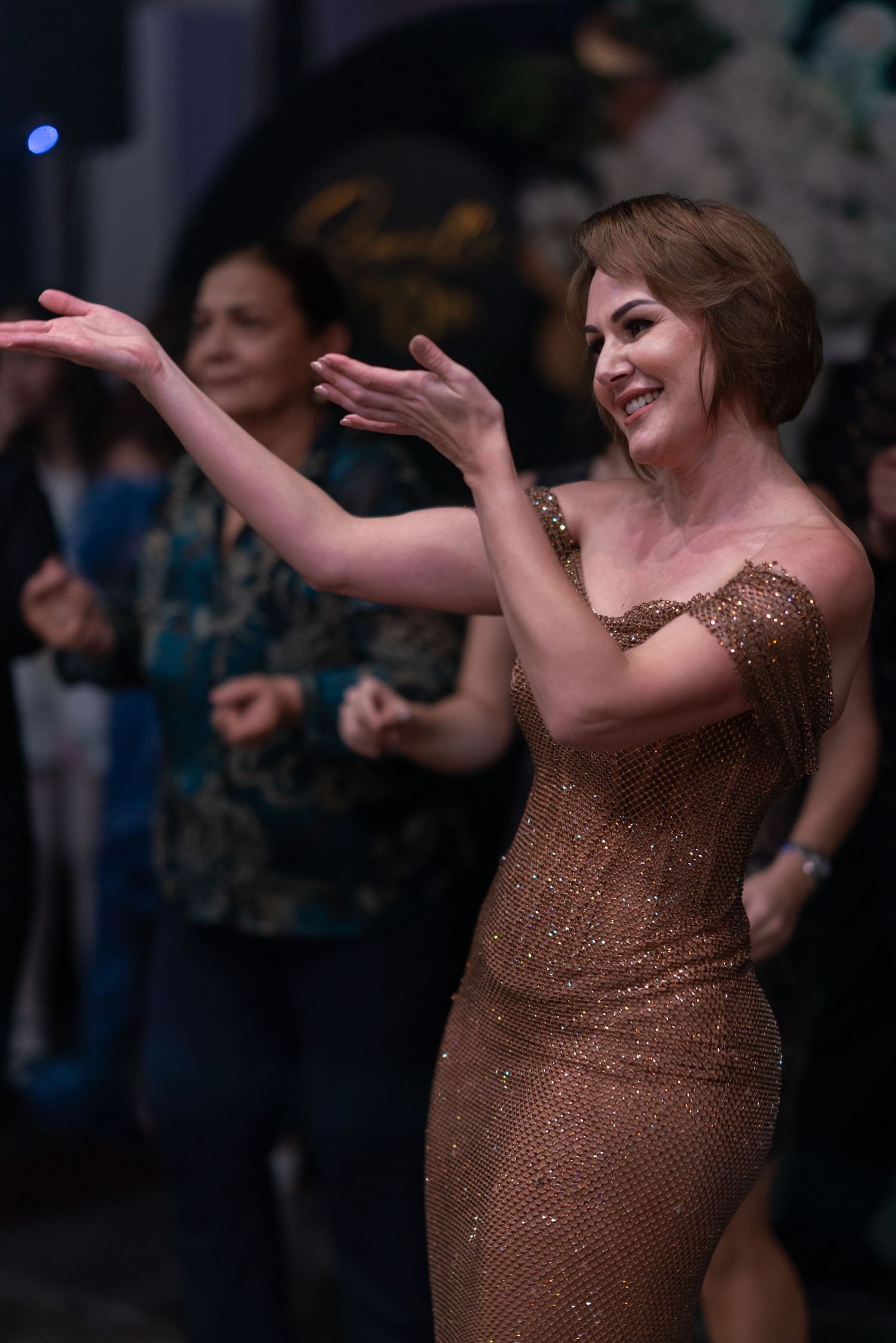 Person in sparkling bronze dress dancing with expressive arm movements on a dark dance floor.