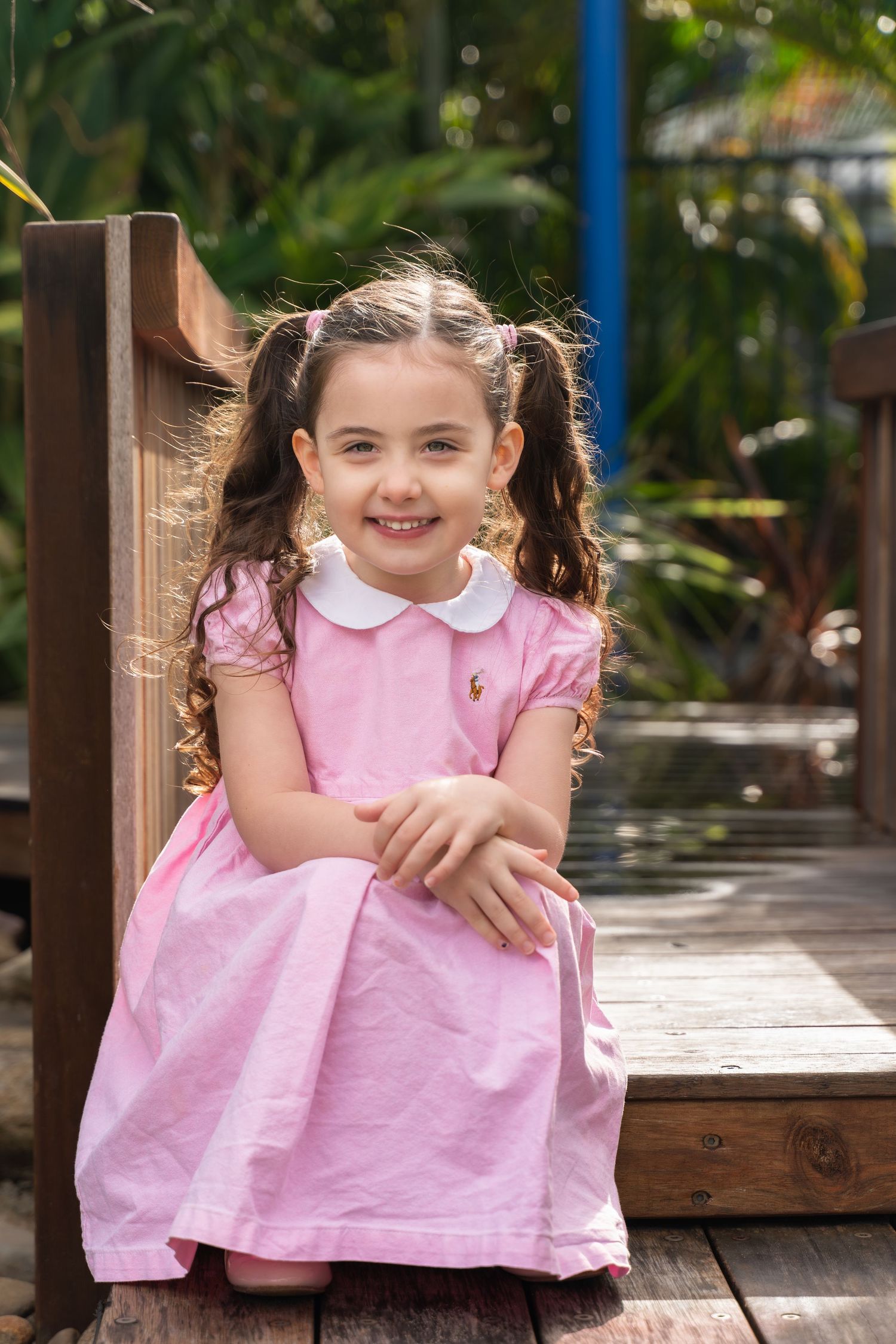 Young girl in pink dress with pigtails sitting on wooden deck surrounded by lush green garden.