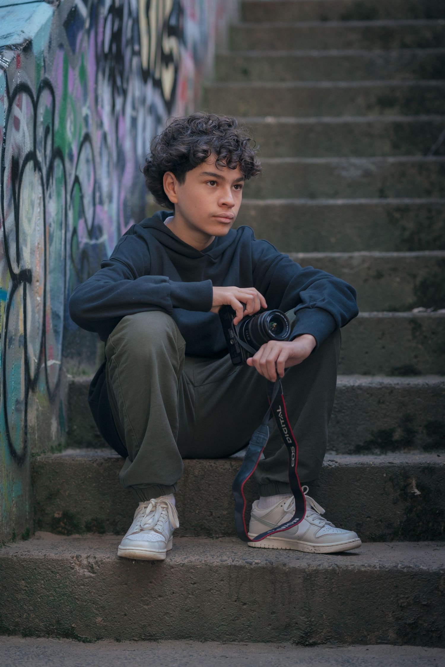Person in casual wear sitting on urban steps with camera near graffiti wall.
