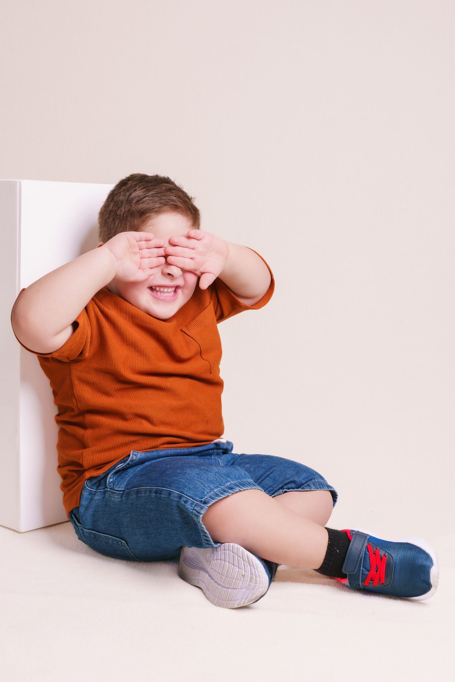 Playful young boy in an orange shirt and denim shorts covering his eyes while sitting against a white cube.