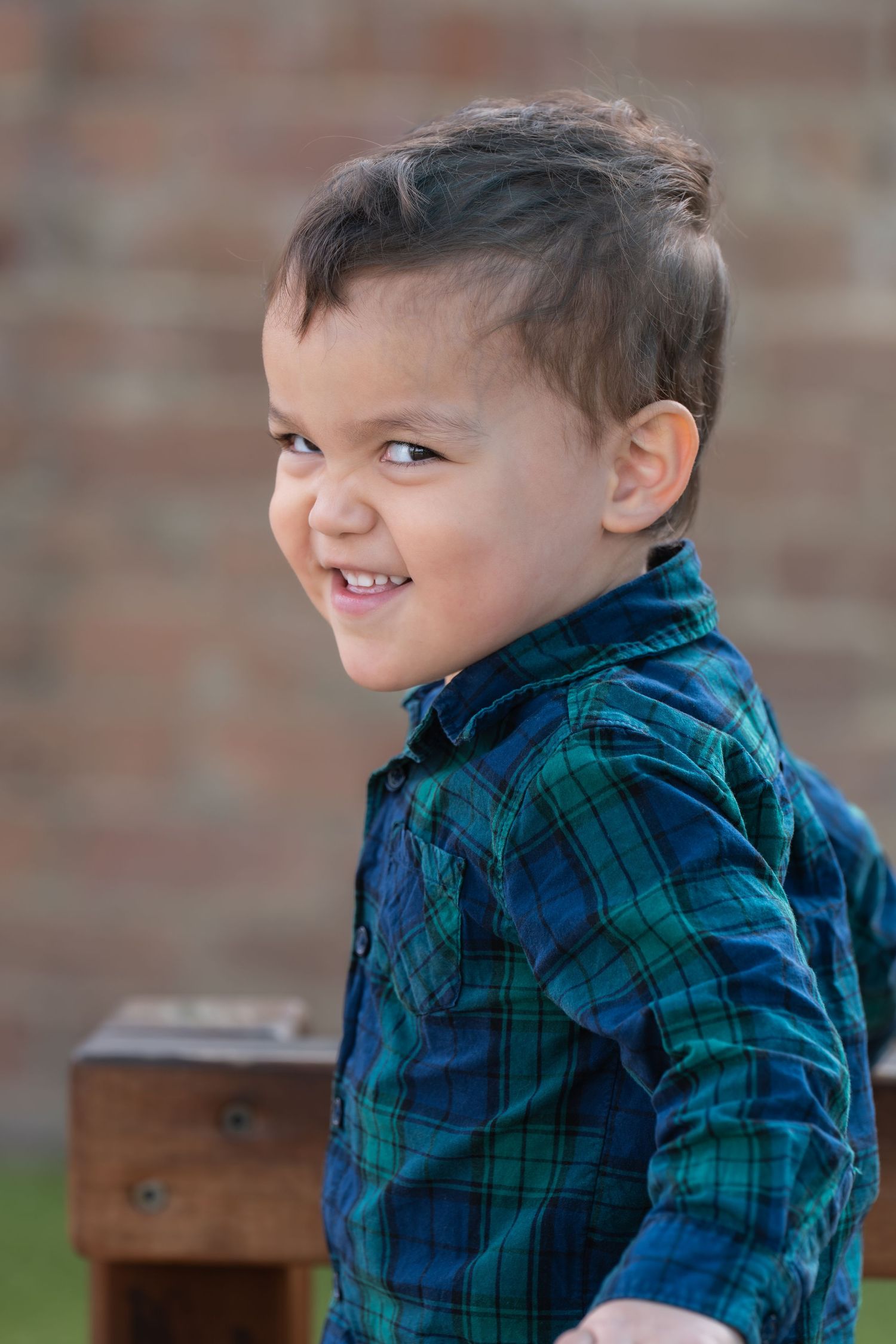 Smiling toddler boy in a blue plaid shirt looking over his shoulder outdoors near a brick wall.