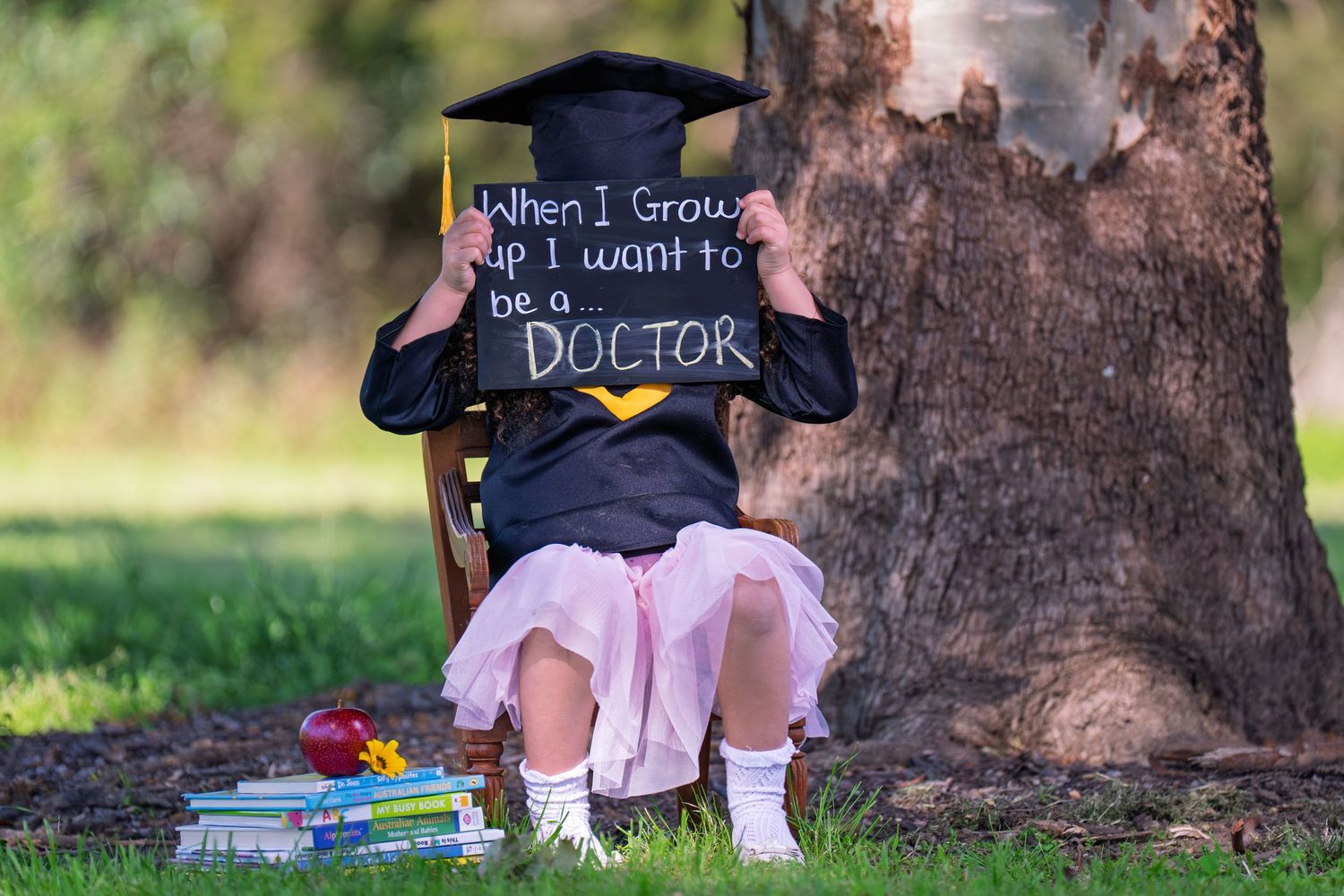 A graduation photo prop and degree books lean against a large tree trunk in an outdoor setting.