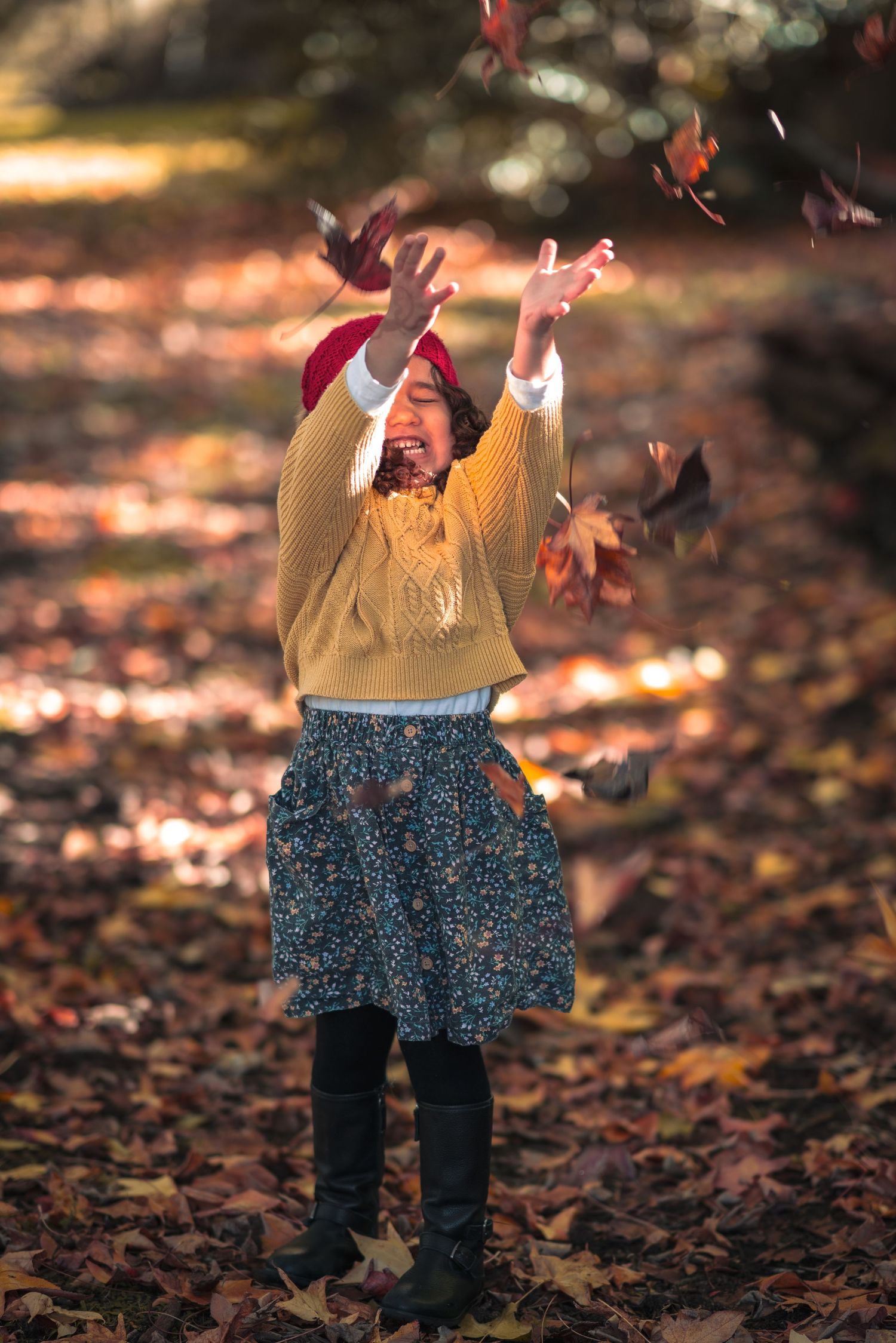 A figure in a yellow sweater and patterned skirt throws autumn leaves in the air with joy.
