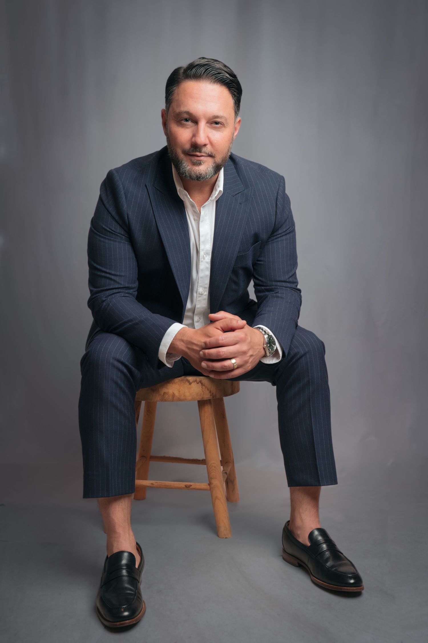 Professional man in navy pinstripe suit and black loafers seated on wooden stool against gray backdrop.