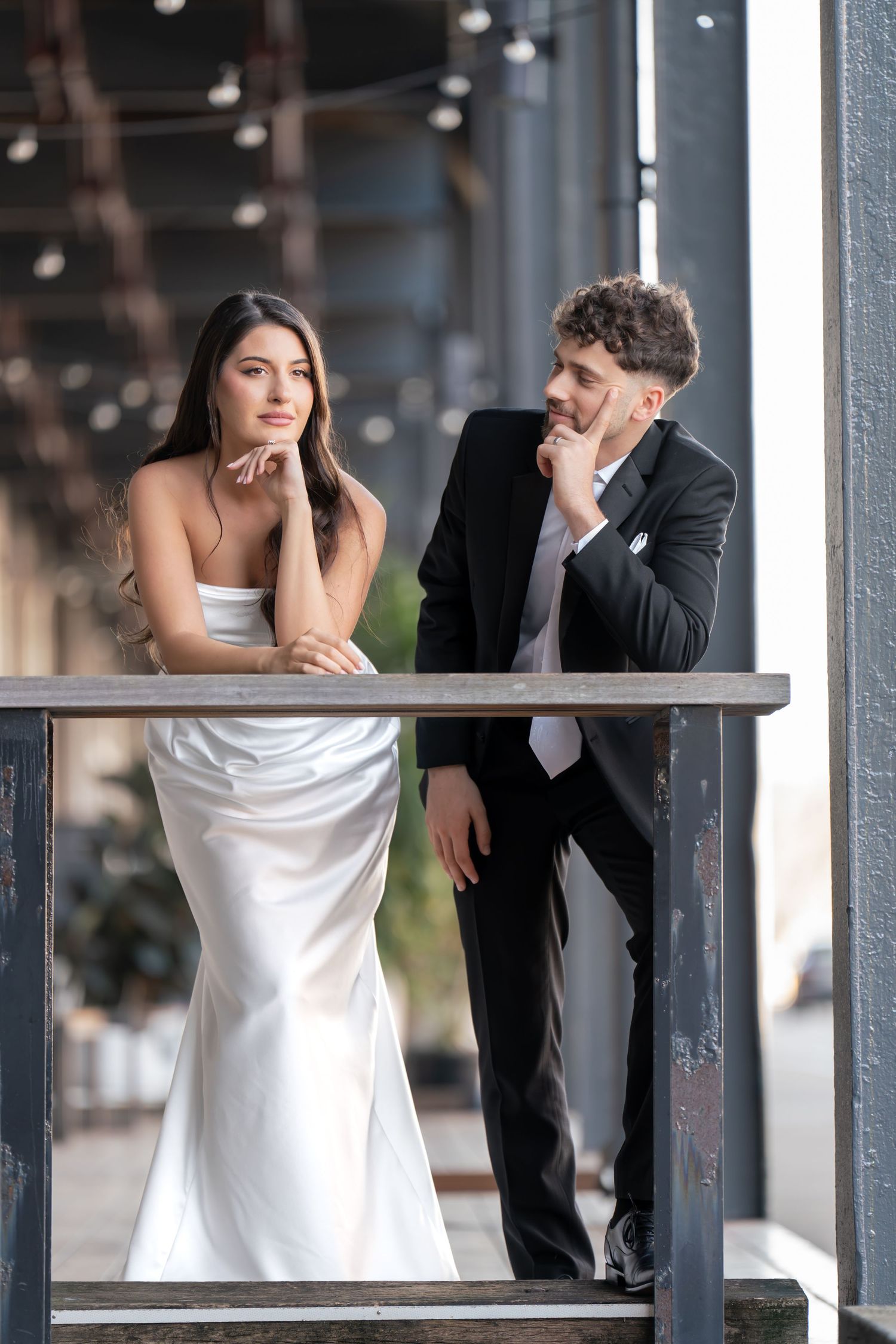 Couple in formal attire leaning on industrial railing in modern setting with dramatic lighting.