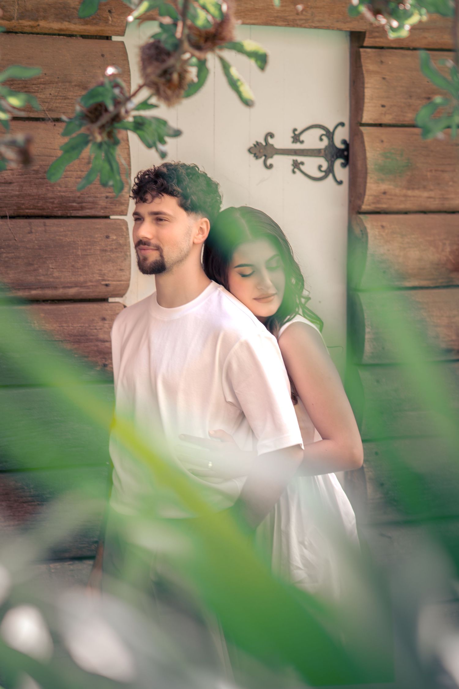 Romantic couple in white outfits share an intimate moment framed by green foliage against a rustic wooden wall.
