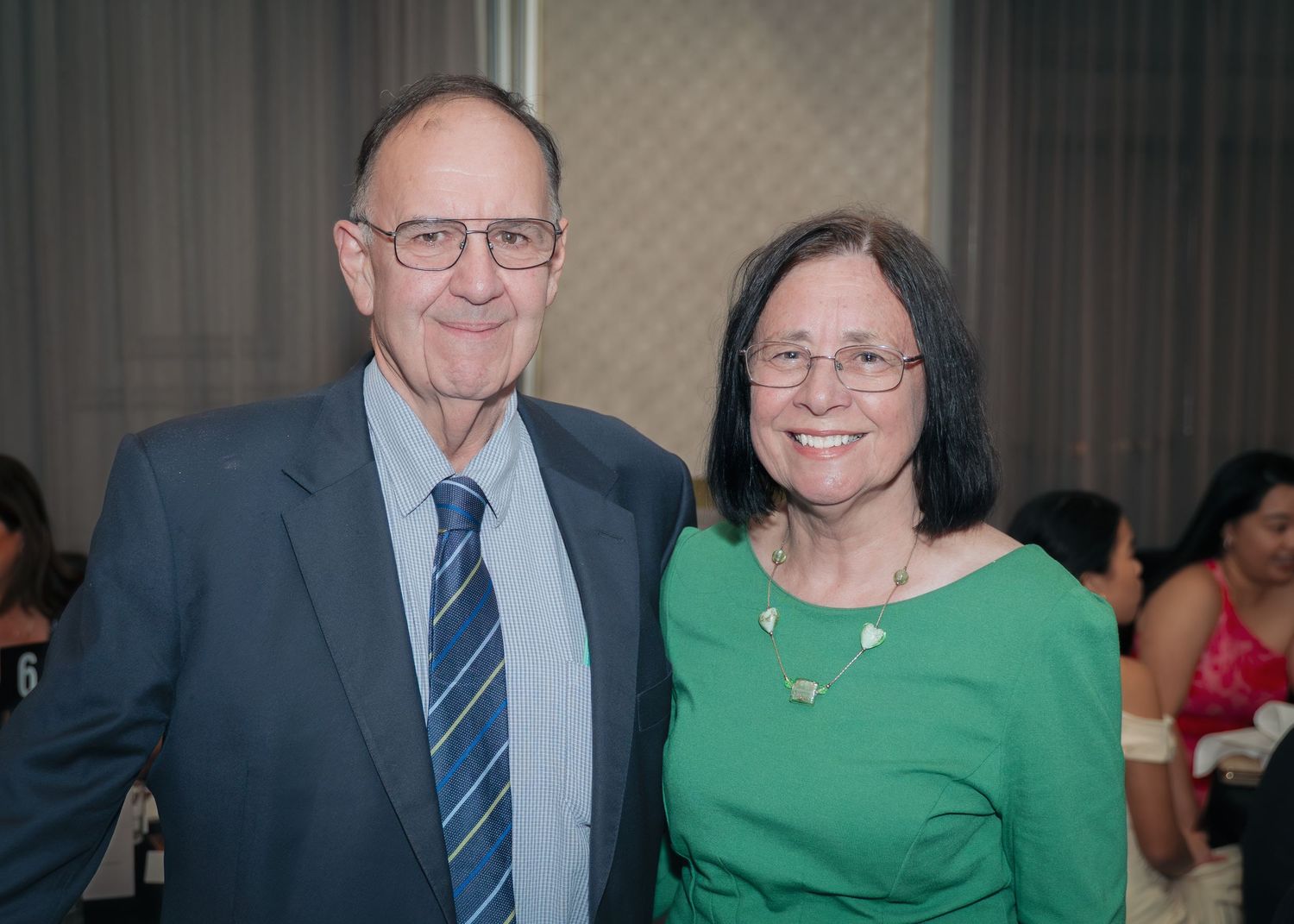 Two people wearing glasses smile together at a formal event, with one in a blue suit and tie and one in a green top.