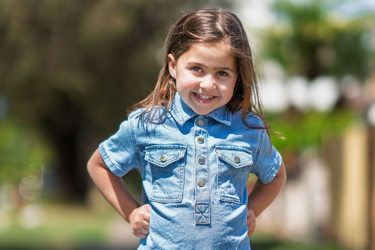 A young child in a denim shirt poses outdoors against a blurred green background.