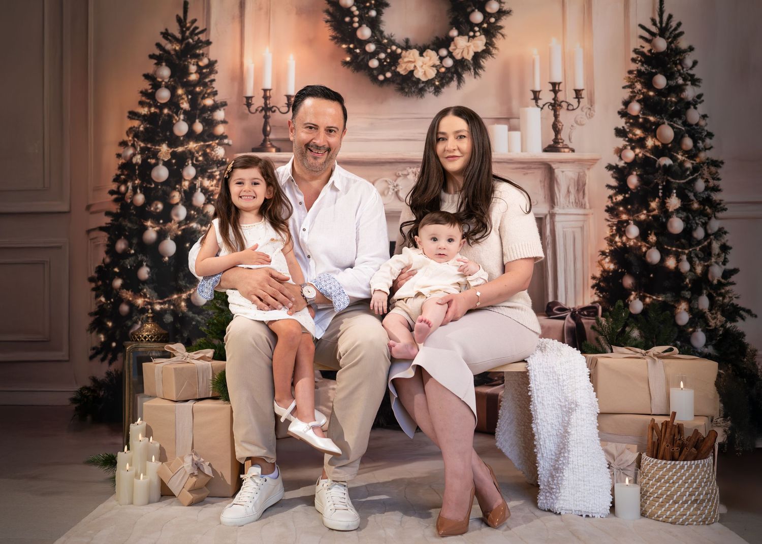 Family wearing white sits together on floor in front of decorated Christmas tree and wreath during holiday photo session.