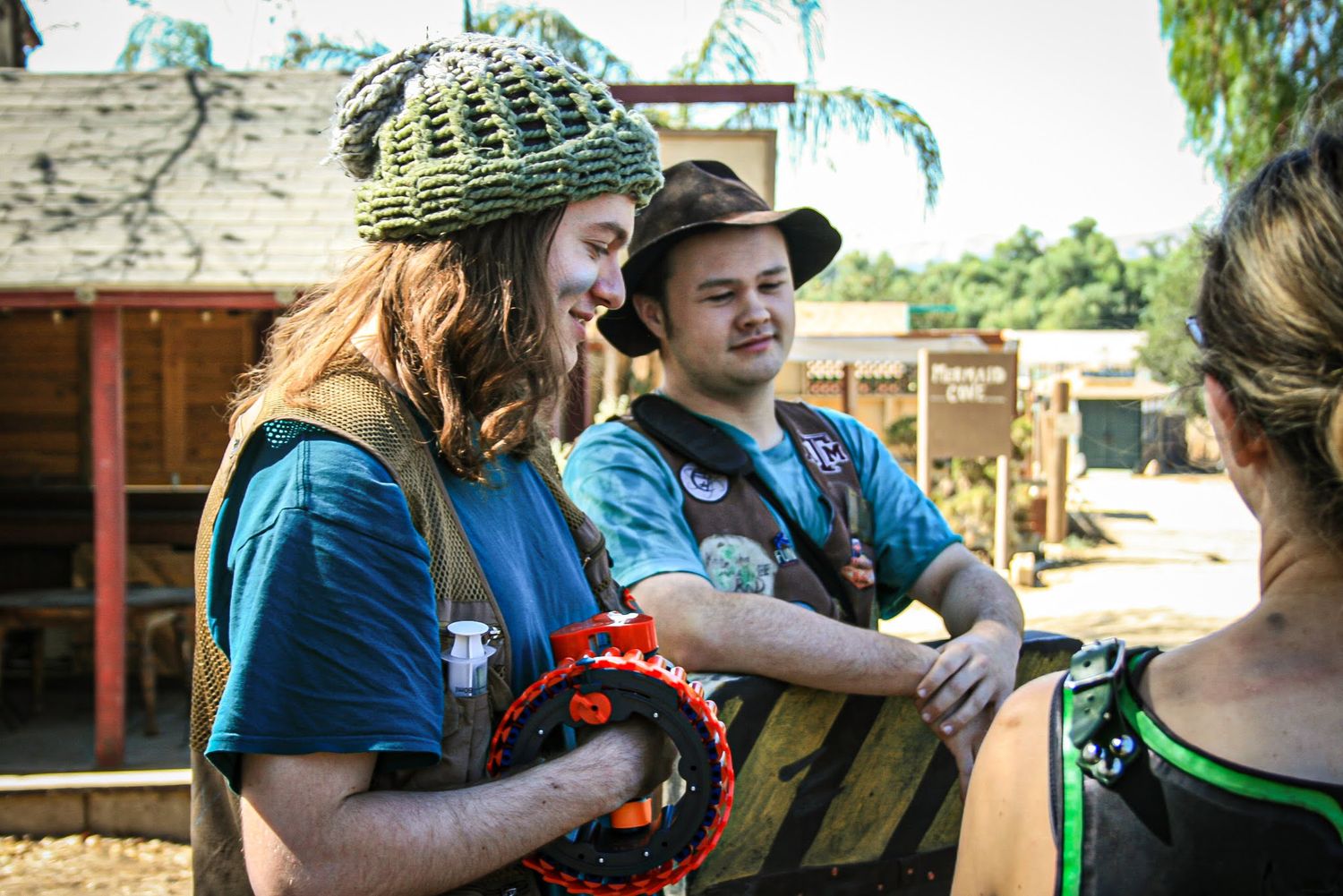 Larper with green hat smiling as he holds a nerf gun, talking with friends.
