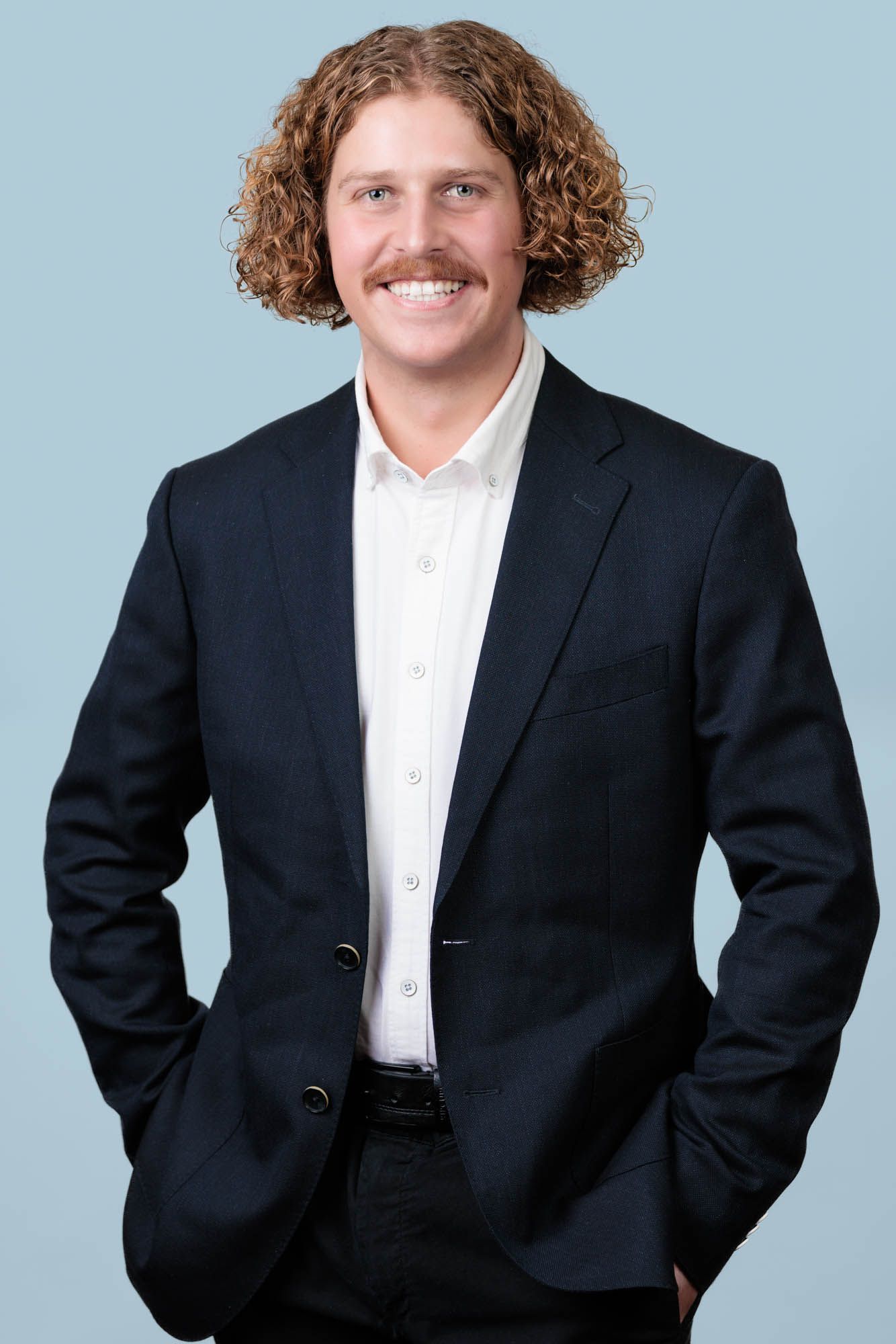 Professional portrait of a young man with curly hair wearing a black suit jacket and white shirt.
