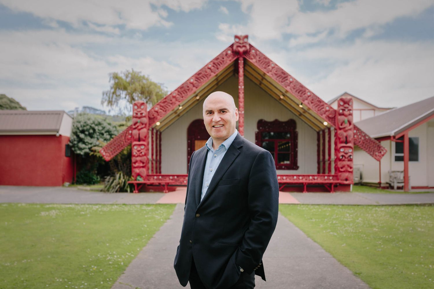 Helmut Modlik standing in front of a traditional Maori meeting house with distinctive carved details and architecture.