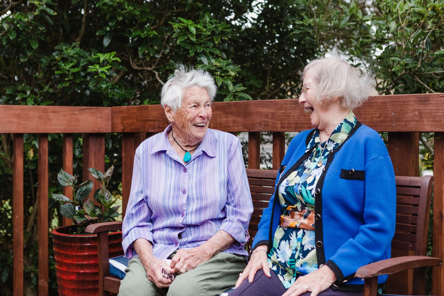 Two elderly friends sharing a conversation while sitting together on a wooden bench in a garden setting.