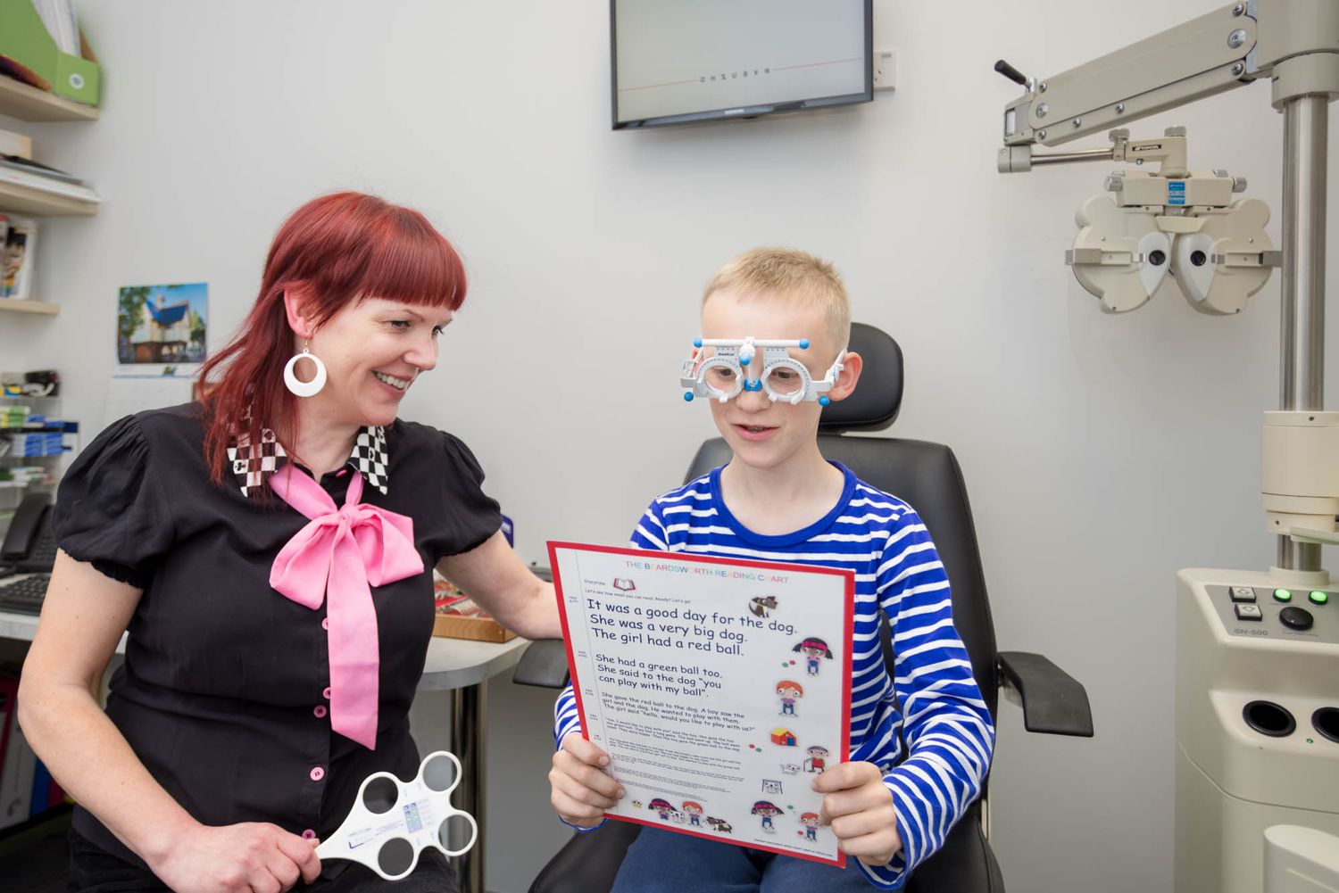 A young patient wearing blue-striped shirt and glasses sits in an exam chair while holding a certificate at an eye doctor's office.