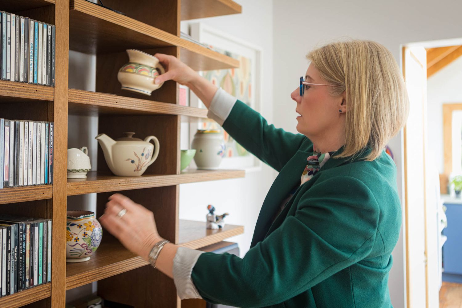 Interior designer in green sweater organising books on wooden wall-mounted shelving units.