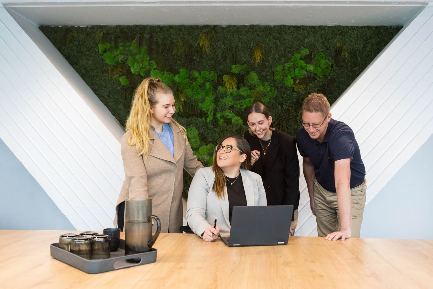 Four colleagues collaborating around a laptop at a modern office desk with a green living wall backdrop.