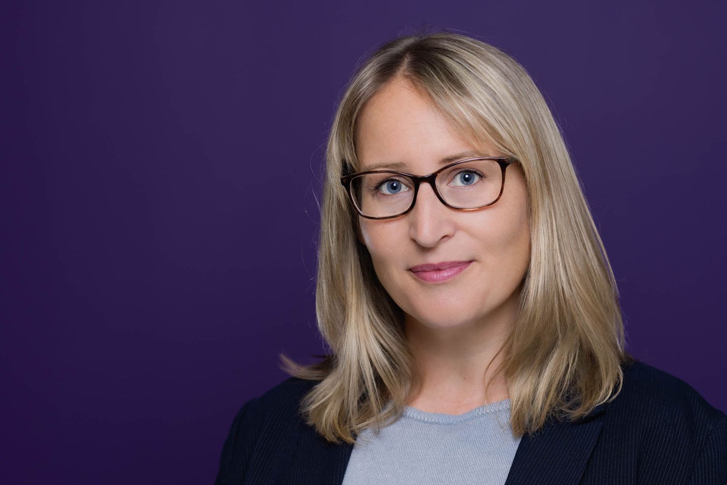 Professional portrait showing a woman with glasses against a deep purple backdrop.