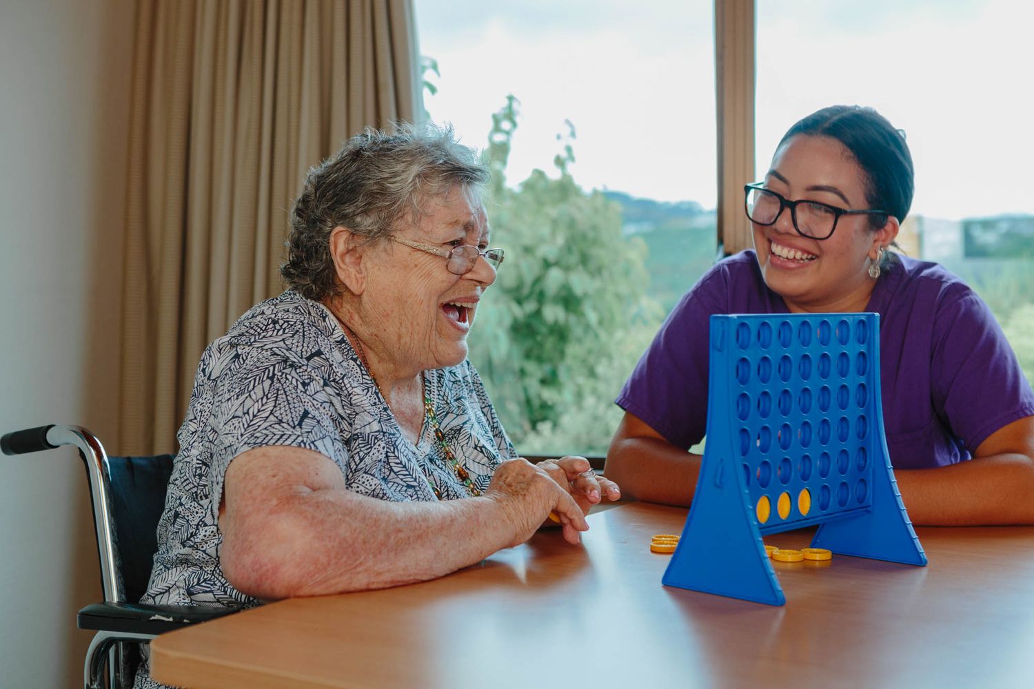 Two people engaged in playing Connect Four game at a table by the window.