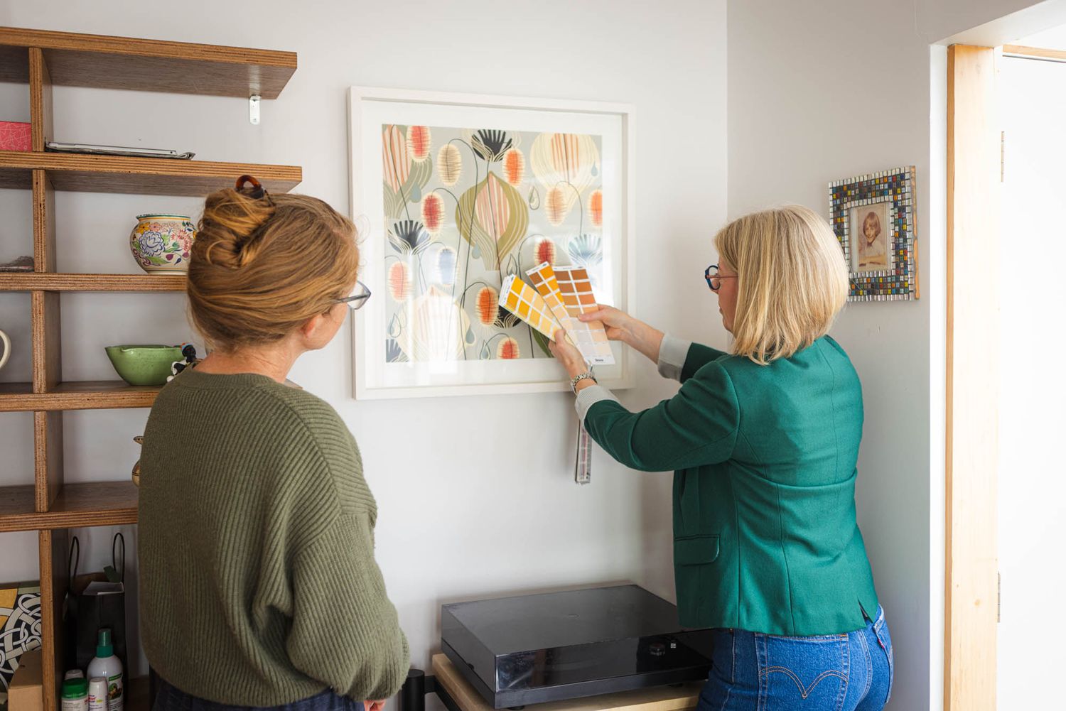 Interior designer arranging photos and artwork on a wooden shelf mounted to a sage green wall.