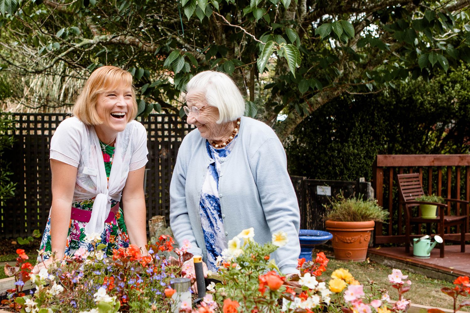 Two people tending to colorful flowers in a garden with potted plants and wooden furniture.