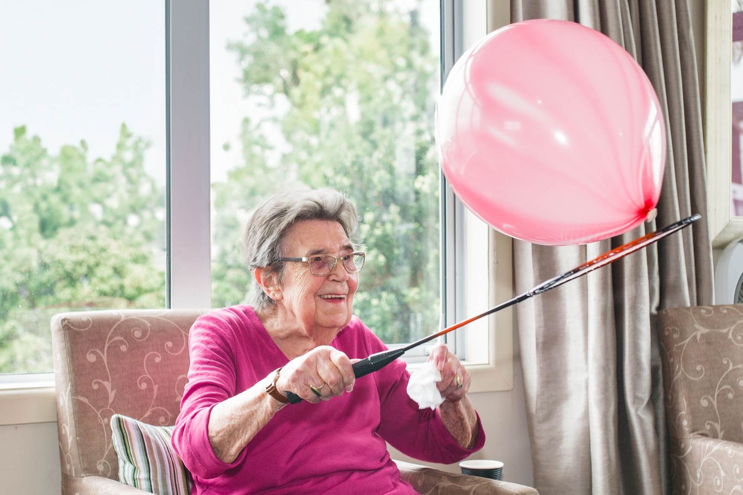 Person in pink sweater holding a pink balloon while sitting in a comfortable chair by a window.