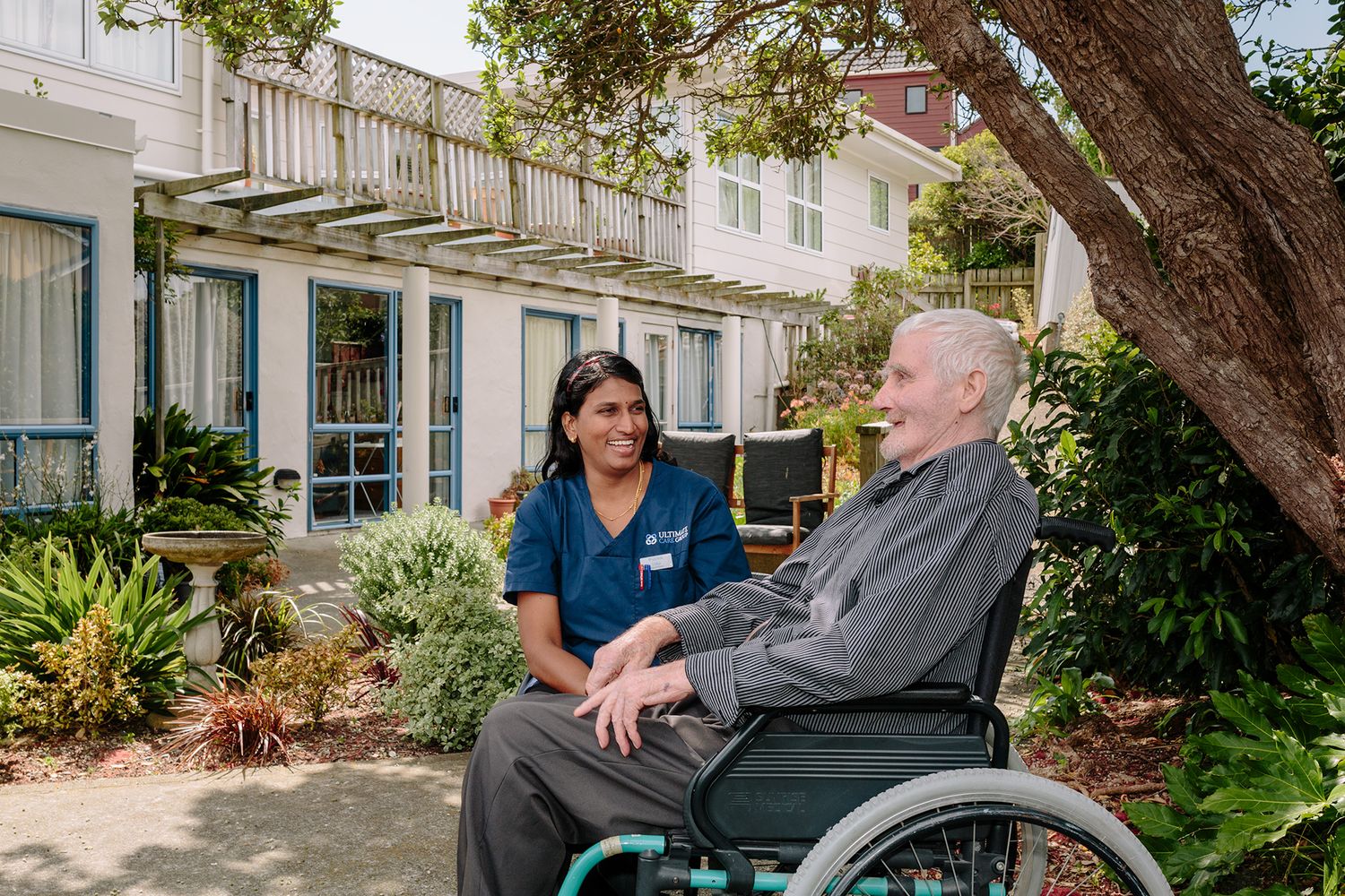 A nurse and an elderly man in a wheelchair share a conversation outside a rest home building.