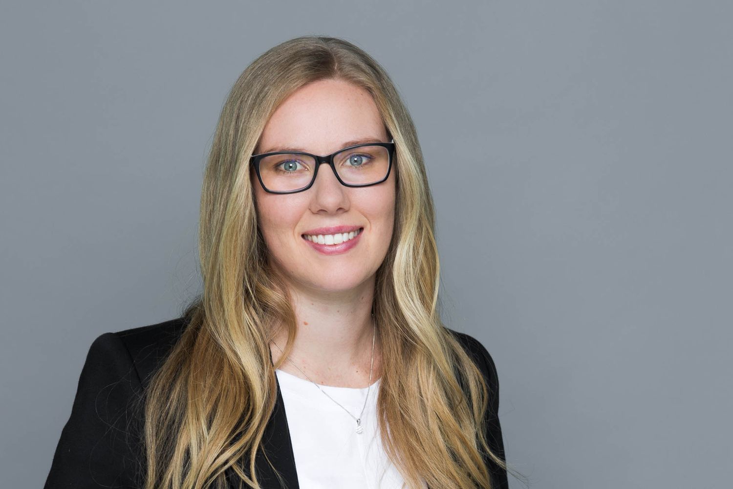 Professional headshot of a smiling blonde woman wearing glasses and a black blazer against a gray background.