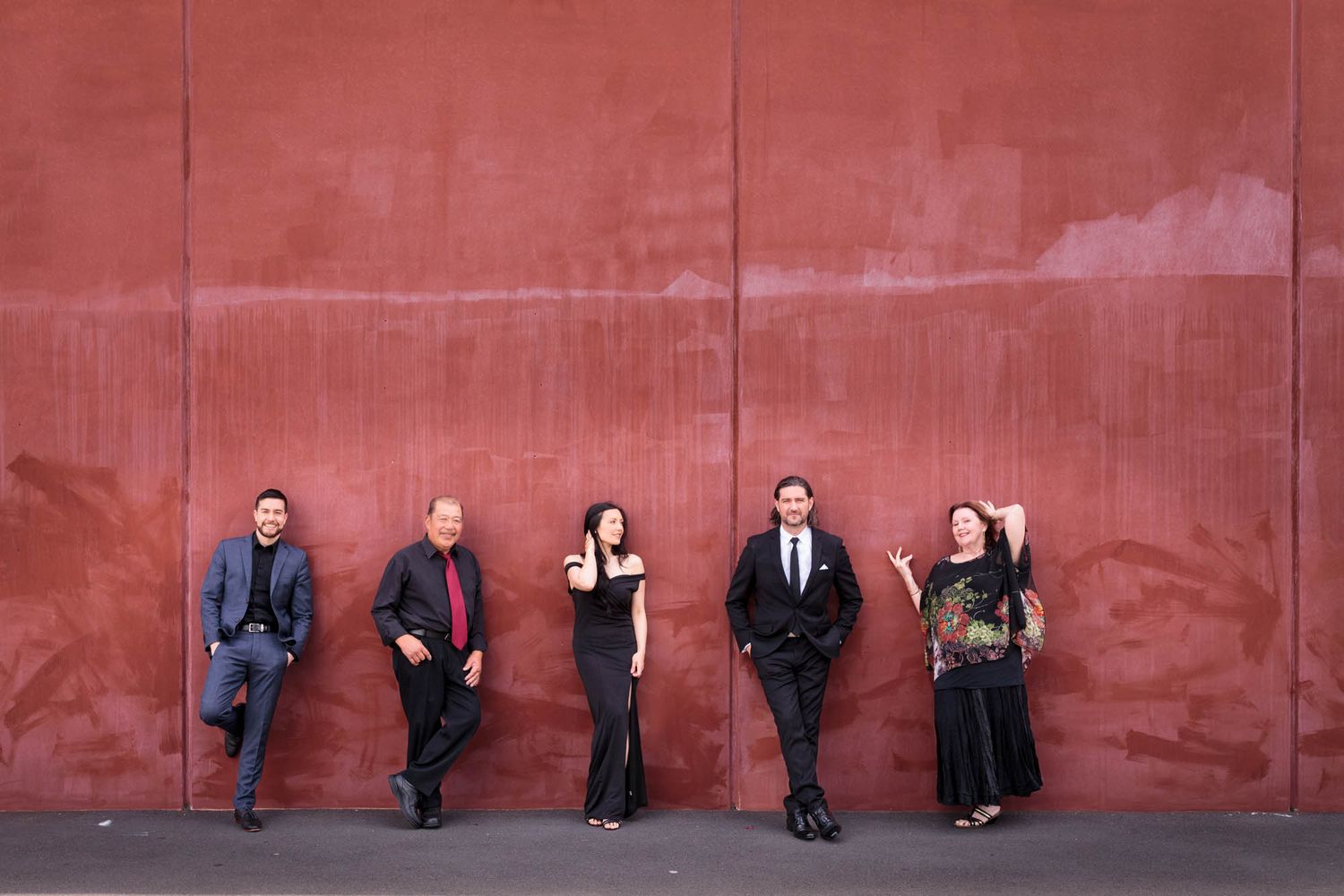 Five people standing next to a red wall wearing dark clothing in an artistic composition.