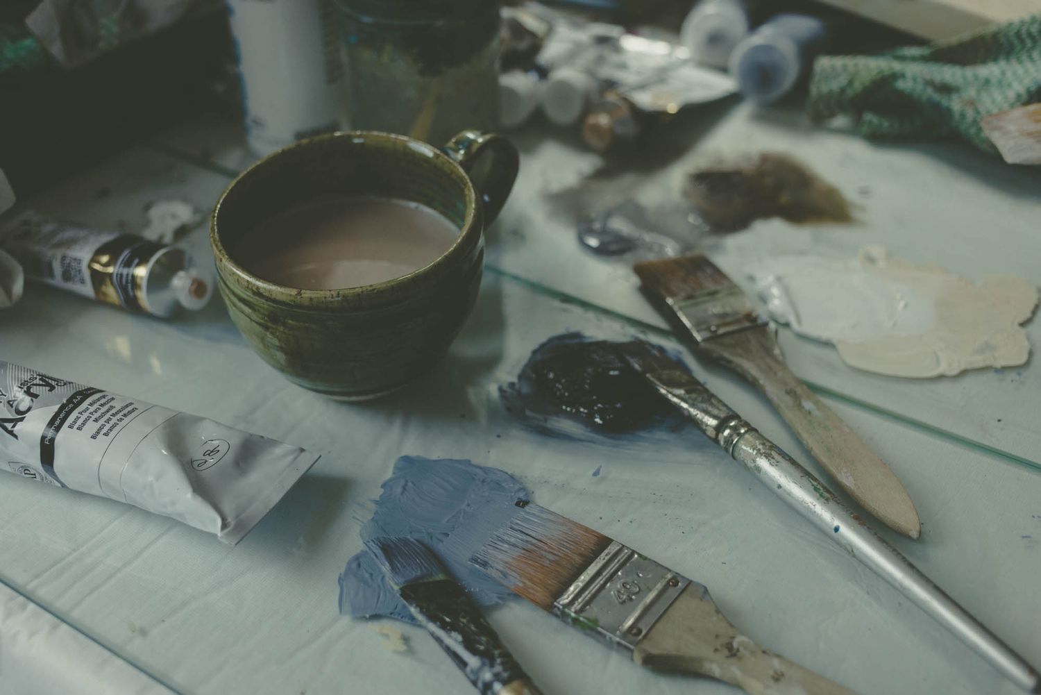 Artist supplies including paint brushes, tubes and a cup on a messy work surface.