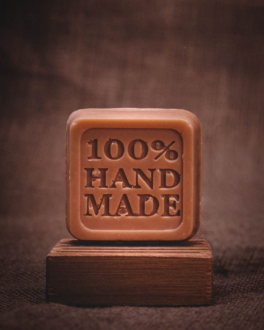 Artisanal soap with embossed text reading '100% Hand Made' displayed on dark textured background.
