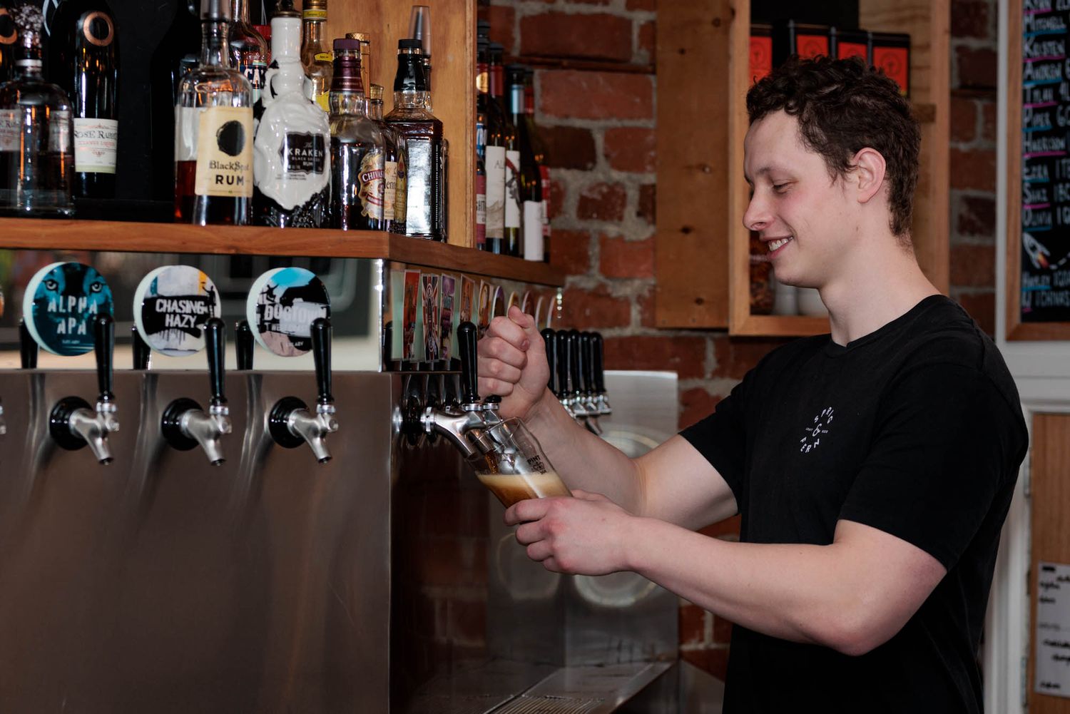 Bartender pouring draft beer from taps at a brick-walled bar counter.
