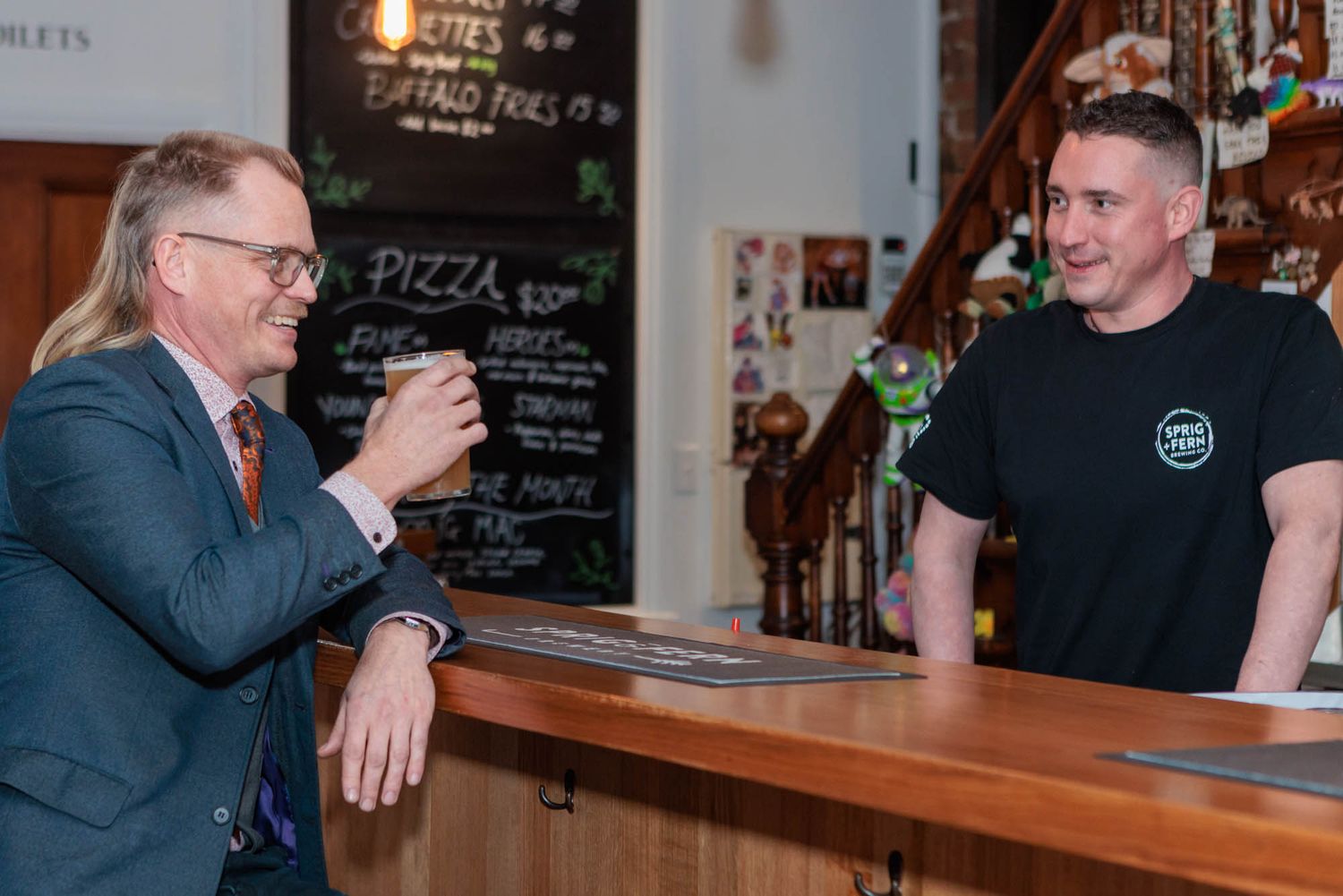 Customer and Sprig and Fern bartender interacting at wooden bar counter with menu board in background.