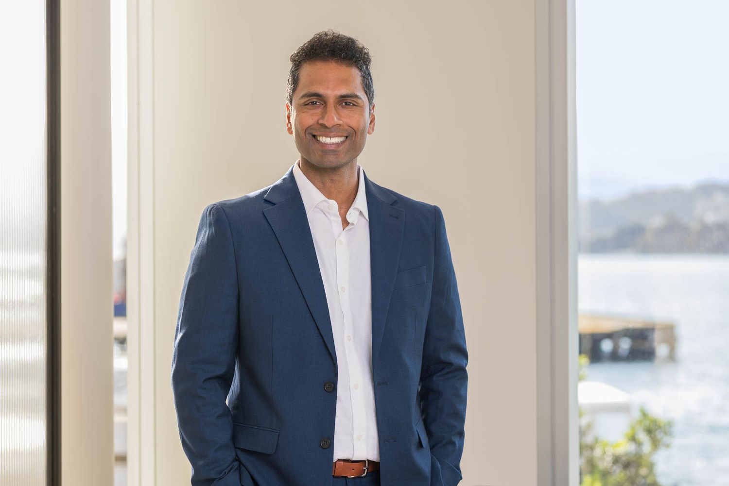 Professional man in blue blazer smiling confidently indoors with waterfront view in background.