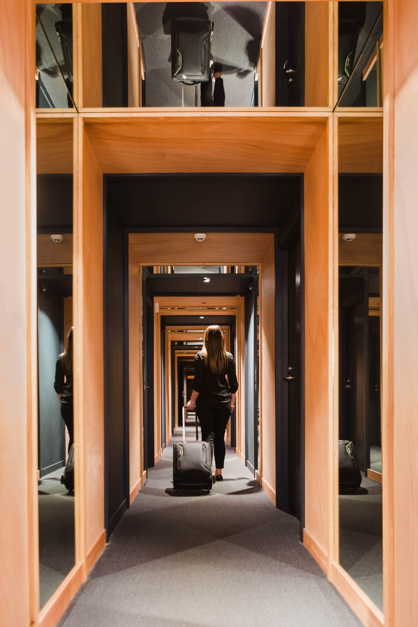 Woman with luggage walking down a modern hotel corridor with wooden frames and mirror panels.